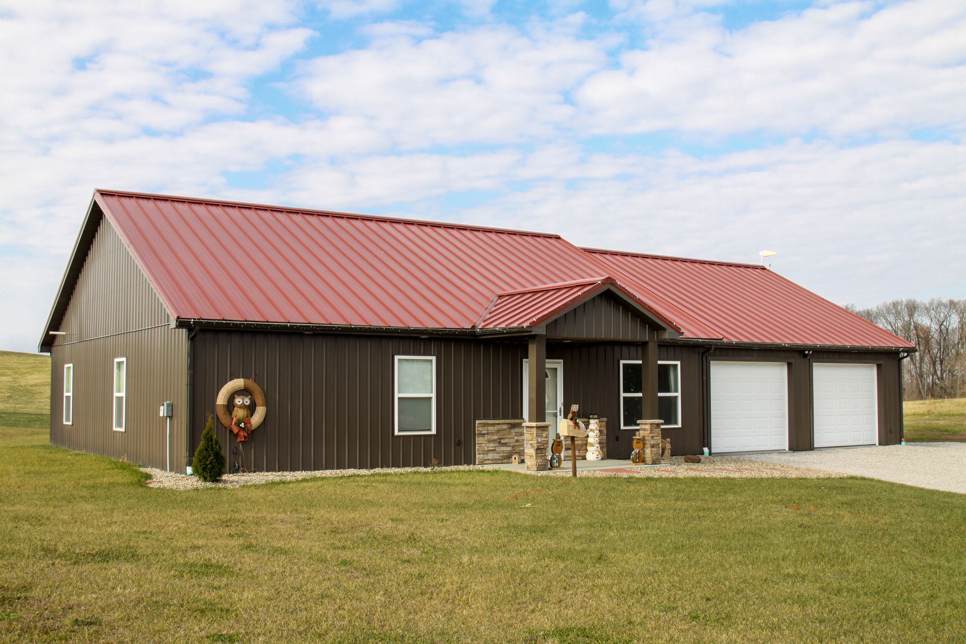 Single-story brown house with red metal roof, front porch, and attached garage on a grassy lot