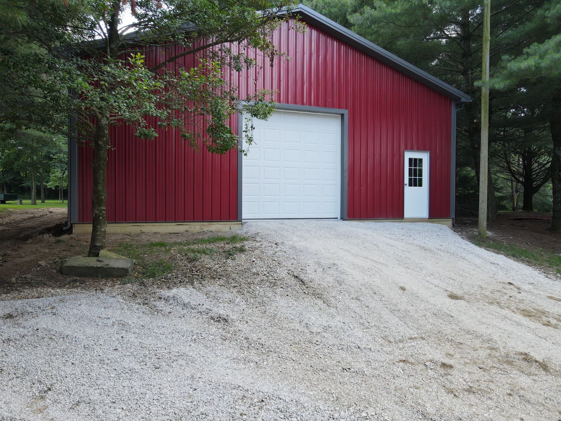 Red metal barn with white doors on a gravel driveway, surrounded by trees.