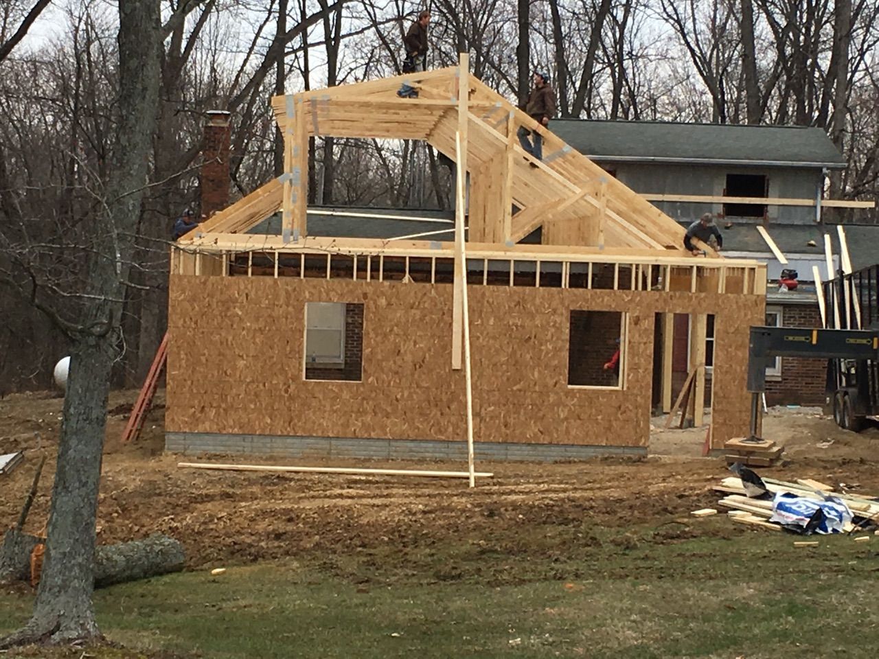 House under construction with exposed wooden framing and plywood walls in a wooded yard