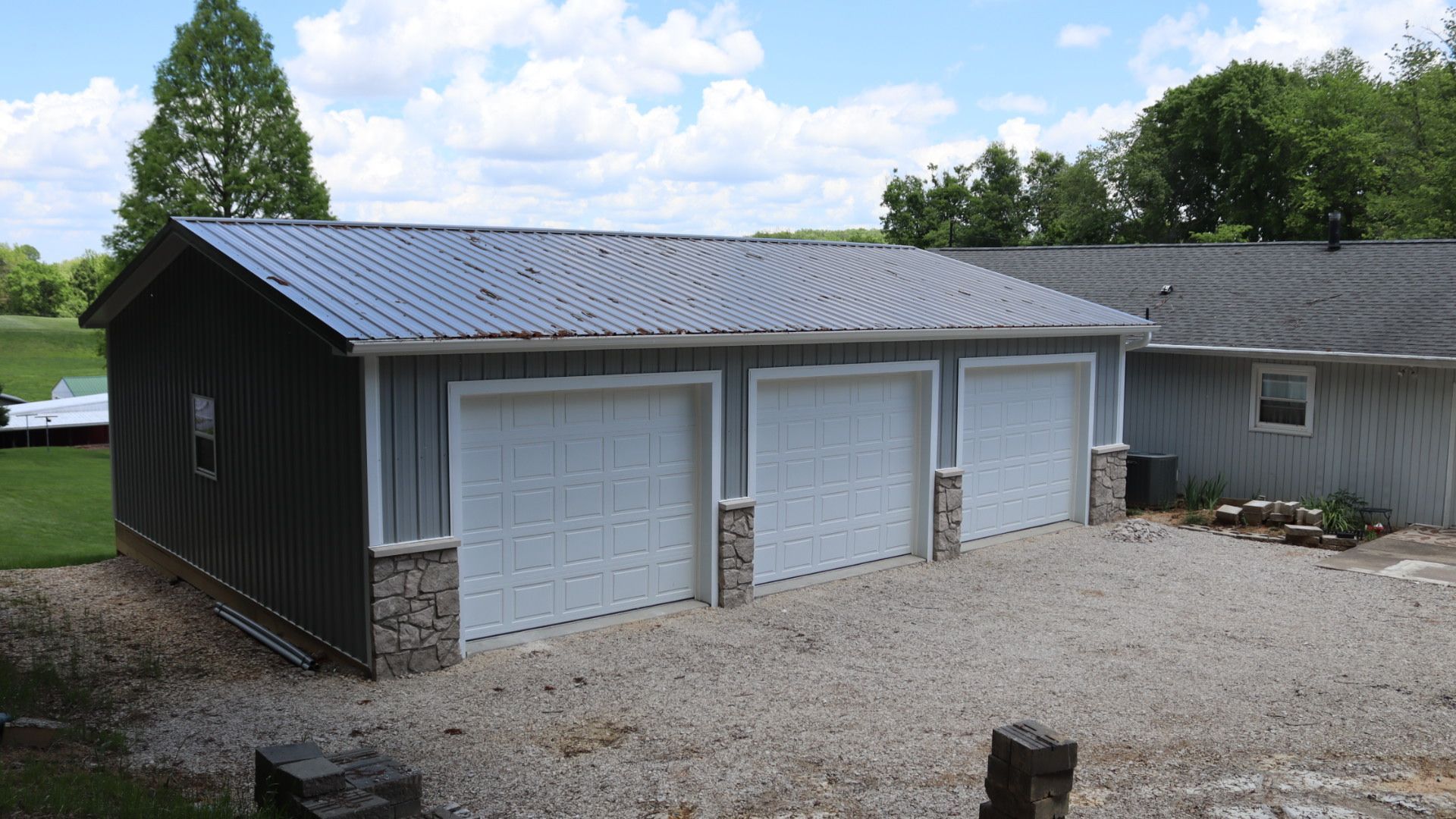 Detached black garage with two white doors and a gravel driveway, set beside a house and trees