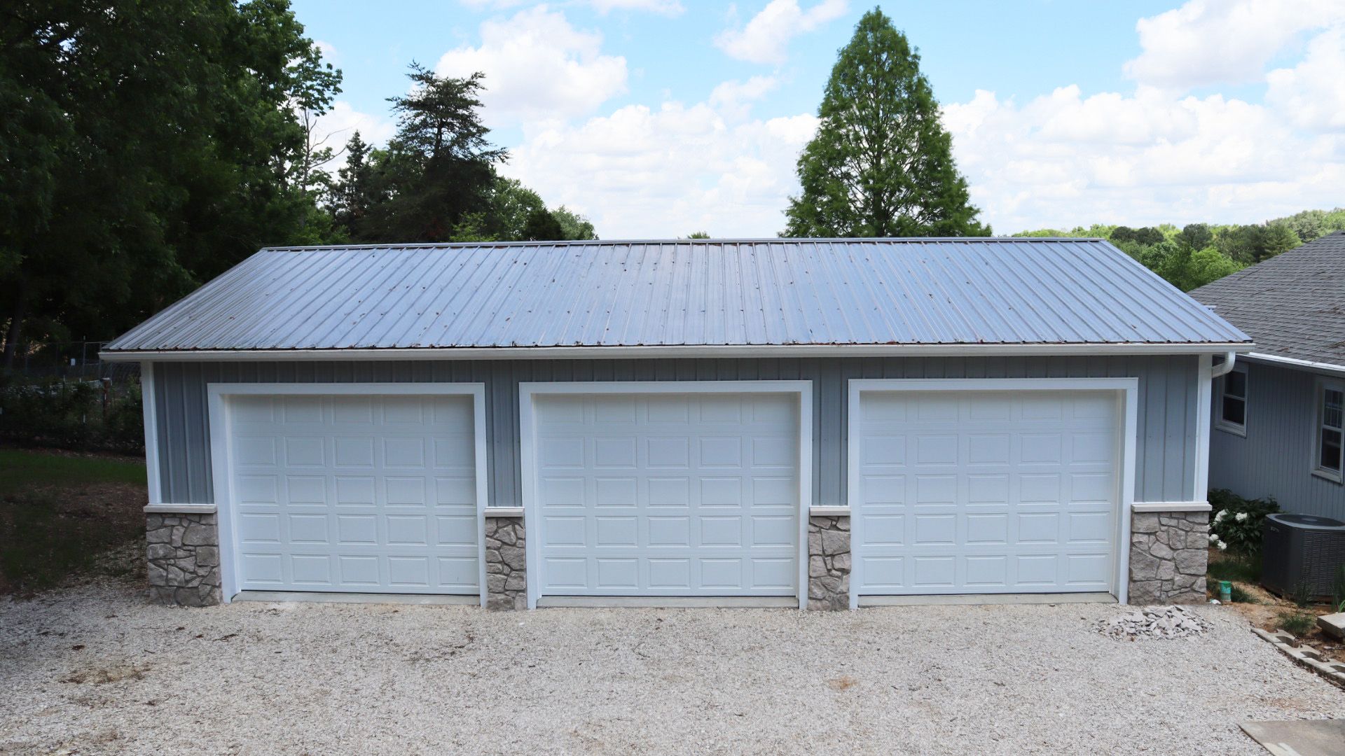 Three-car garage with white doors and gray roof, set on a gravel driveway with trees behind.