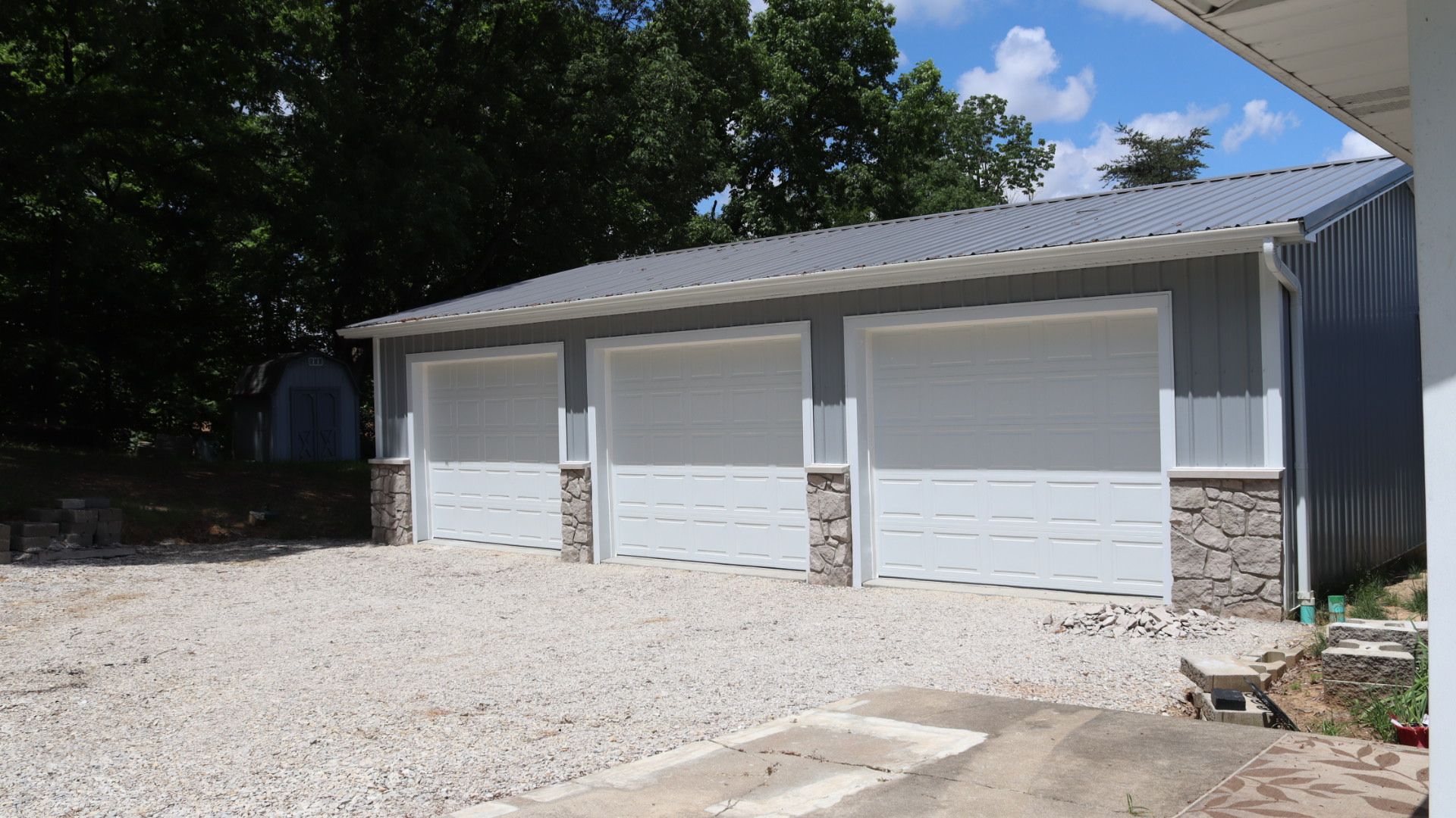 Three white garage doors on a light gray building with a gravel driveway and trees nearby