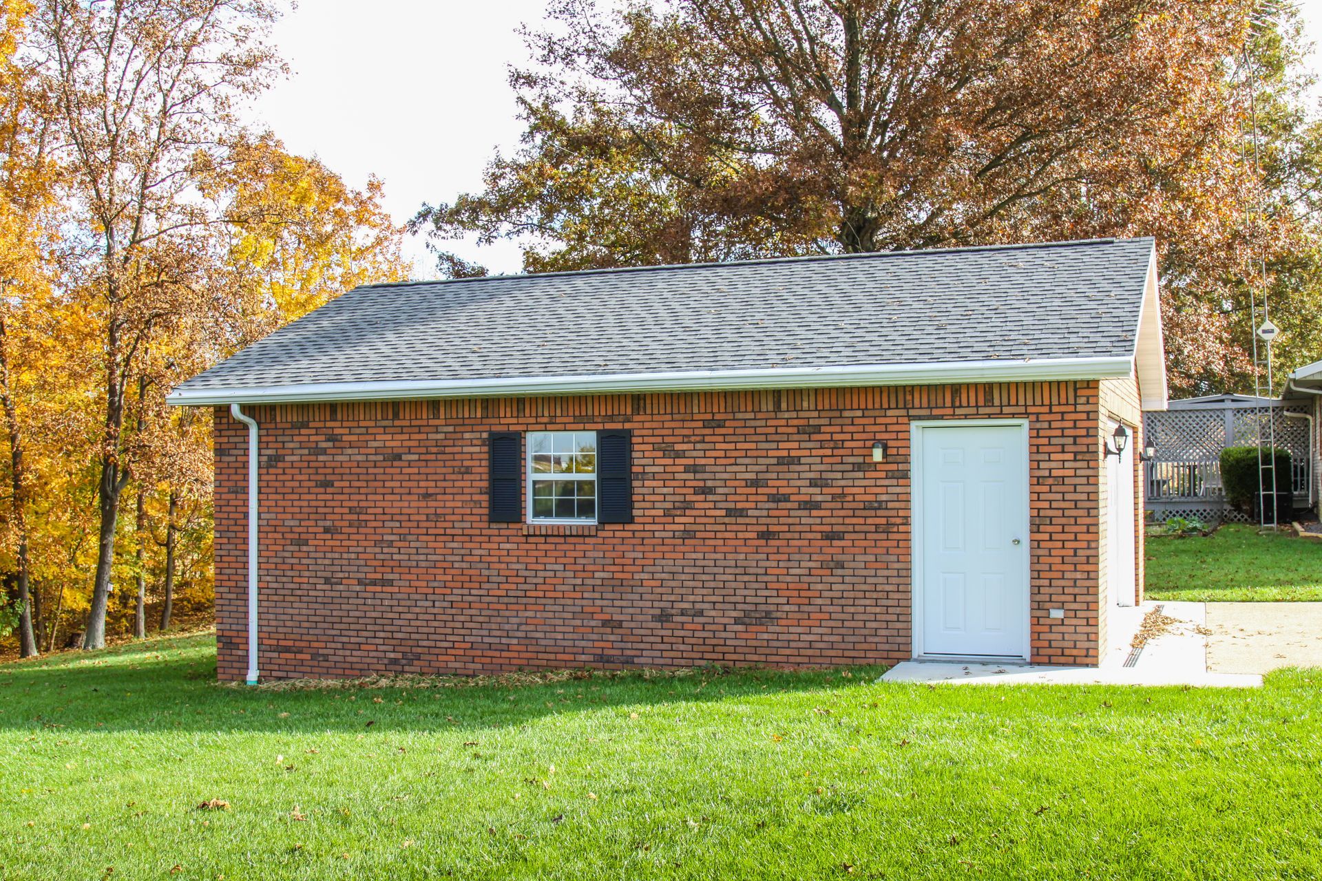 Small red brick shed with a white door and window on a grassy lawn, surrounded by autumn trees