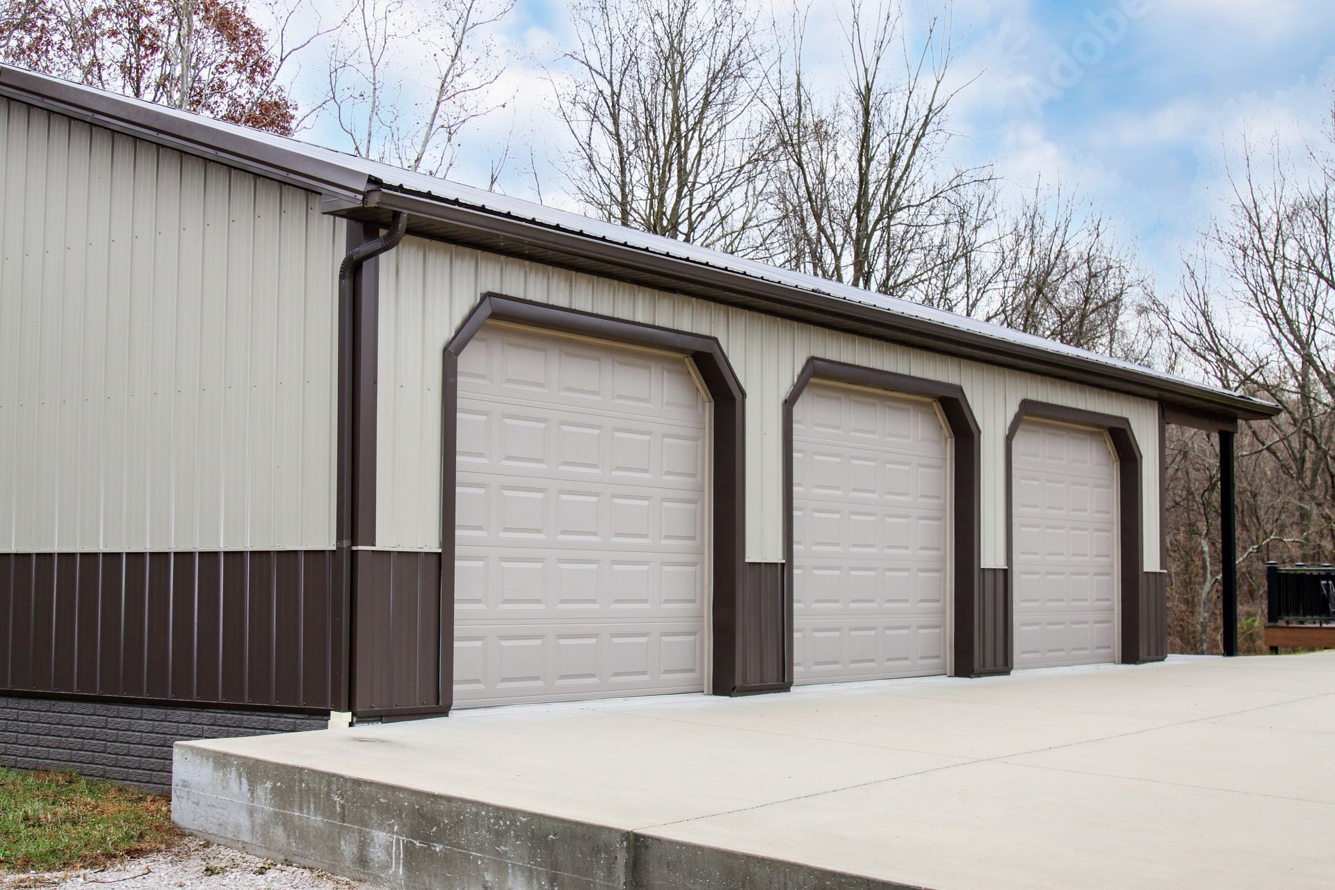 Three white garage doors on a brown metal-sided building with a concrete driveway and leafless trees.