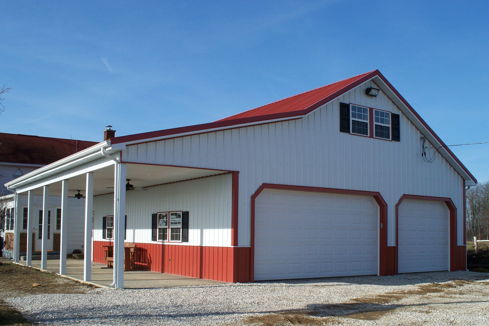White barn-style building with red roof and trim, garage doors, and a covered porch on a gravel lot