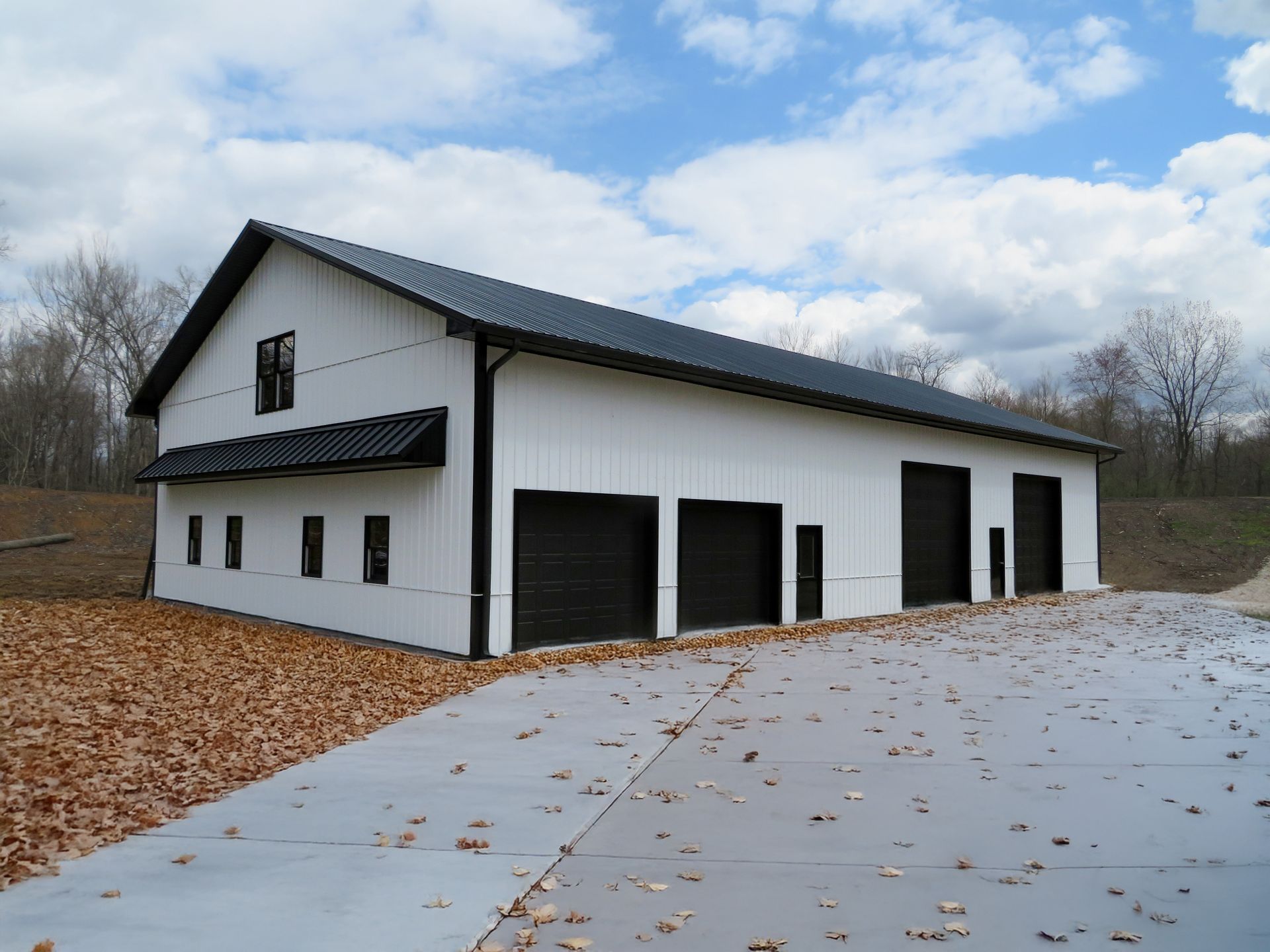 White barn-style building with black trim and garage doors beside a concrete driveway under a cloudy sky