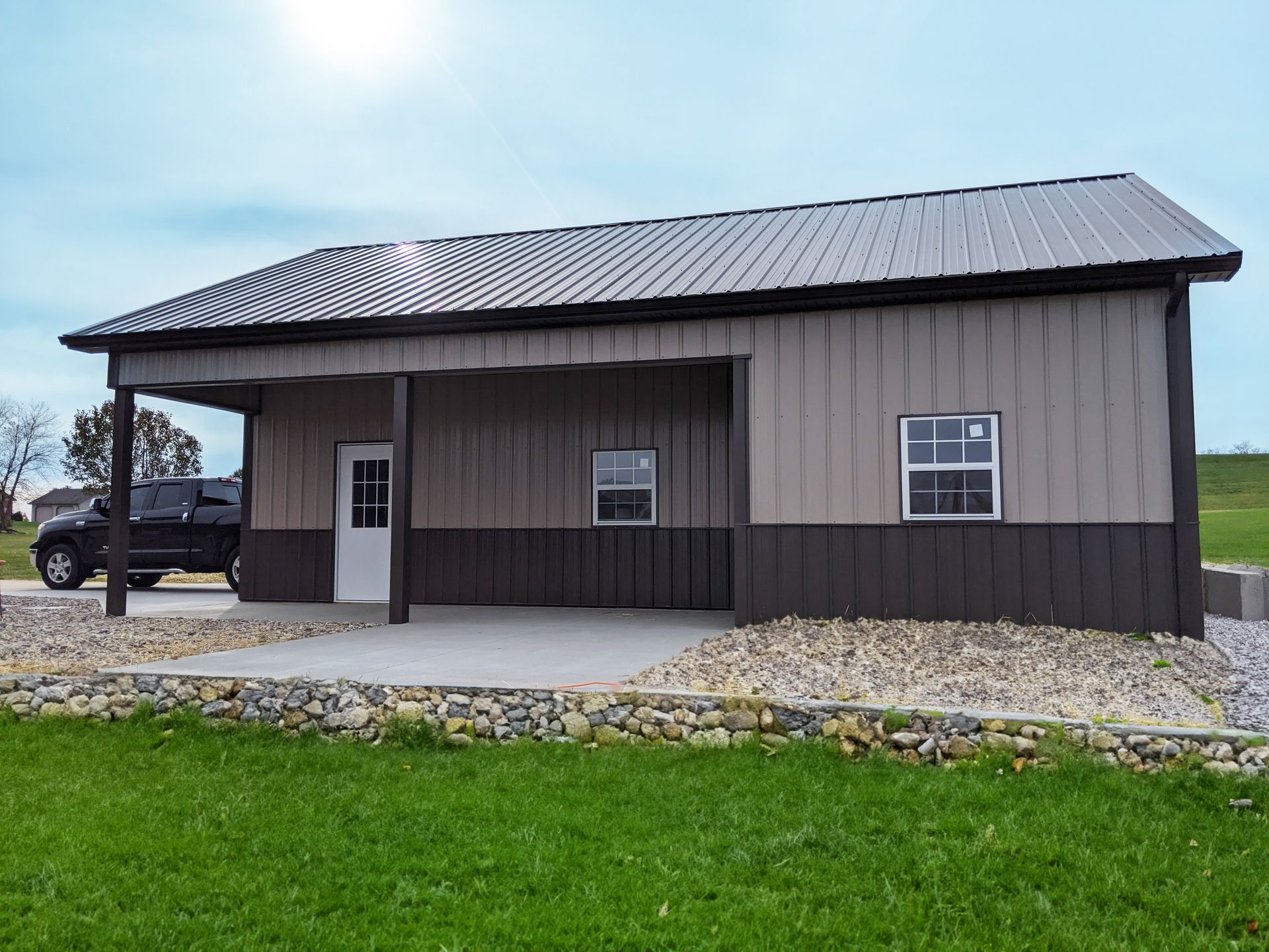 Small gray metal building with a sloped roof, porch, and gravel driveway beside a grassy lawn.