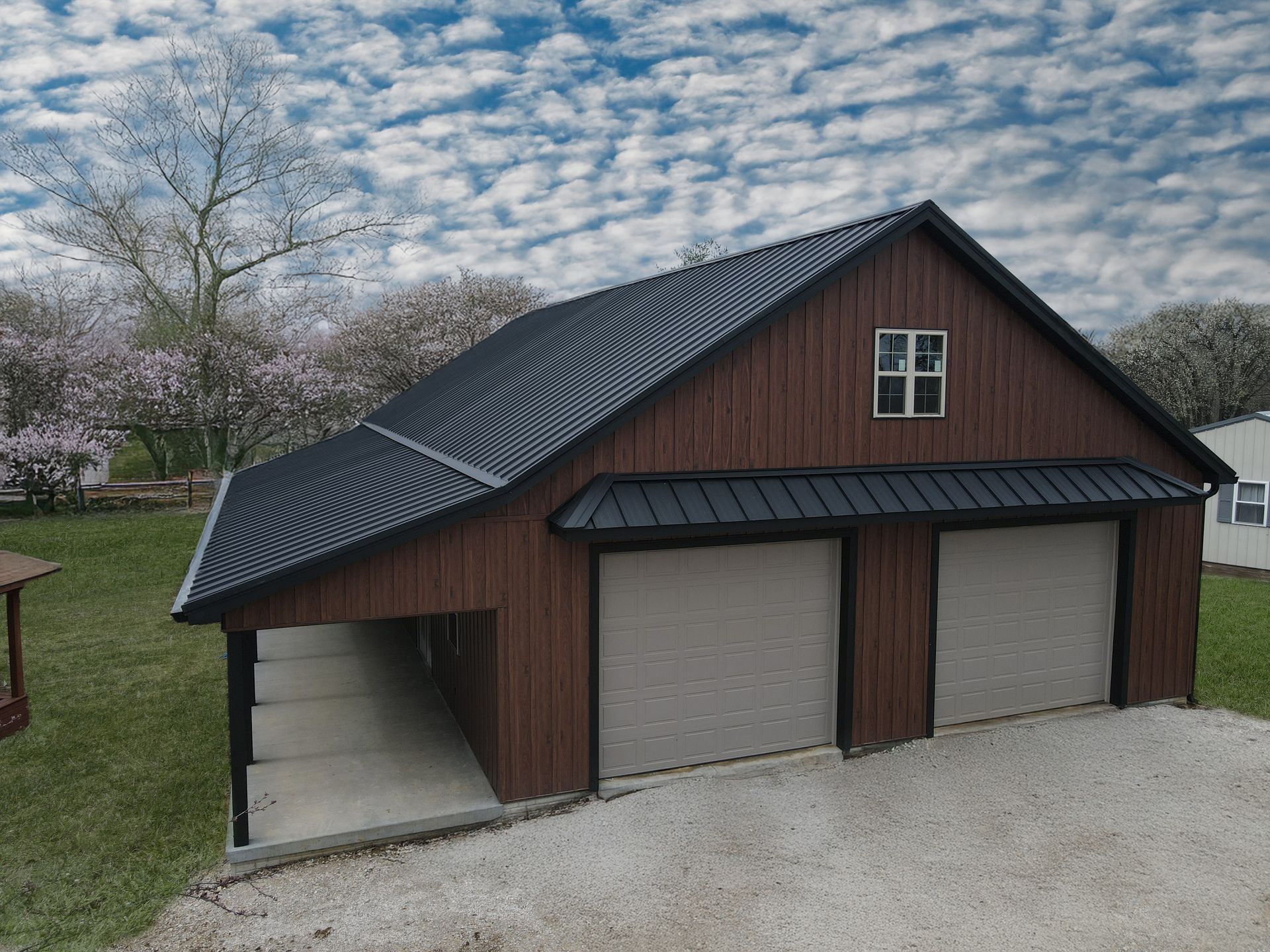 Three-car detached garage with brown siding, black roof, and light gray doors in a yard with trees and gravel drive