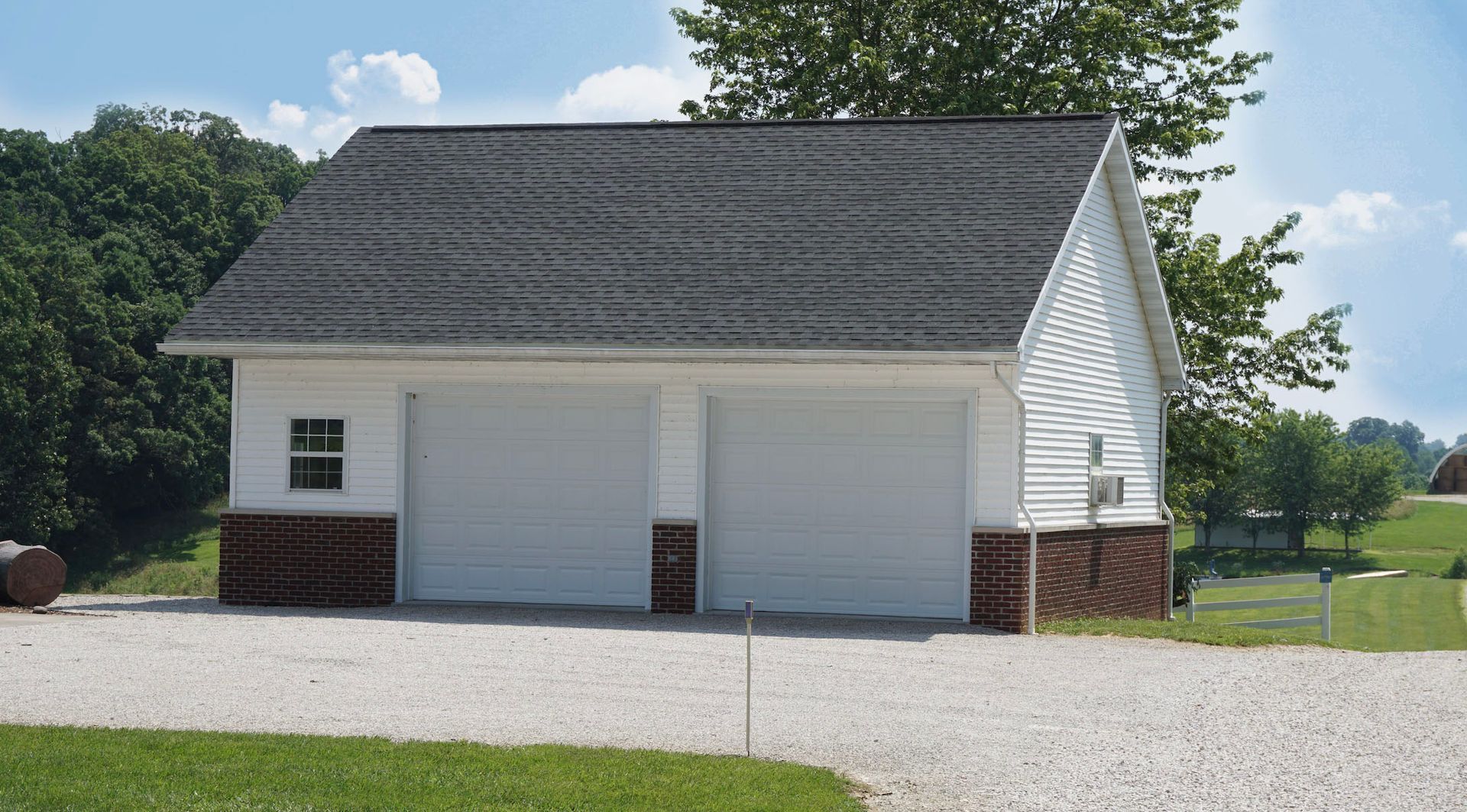 White two-car garage with gray roof and brick base on a gravel driveway, surrounded by grass and trees