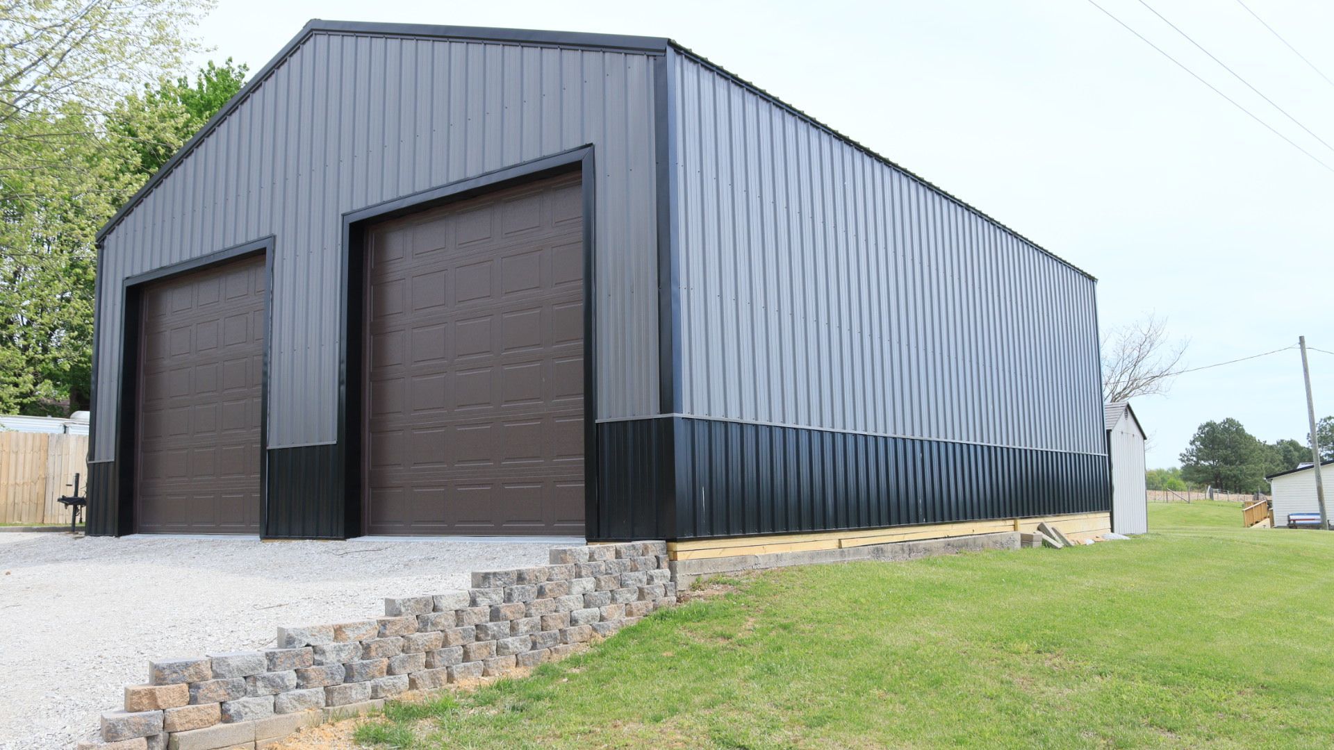 Dark gray metal warehouse with two large garage doors beside a sloped grassy lot