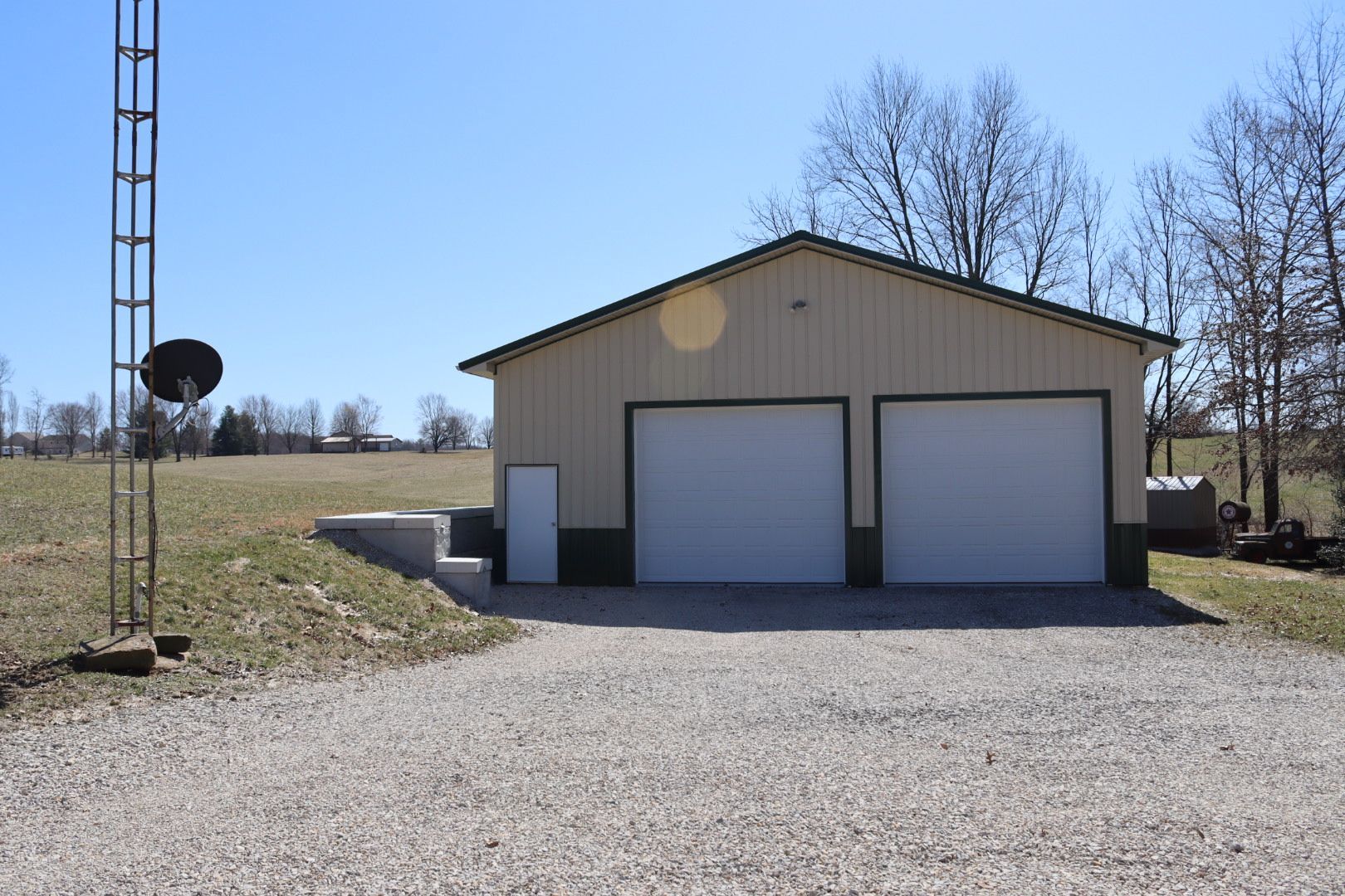 Beige garage with two white doors on a gravel driveway in a rural area under a clear blue sky