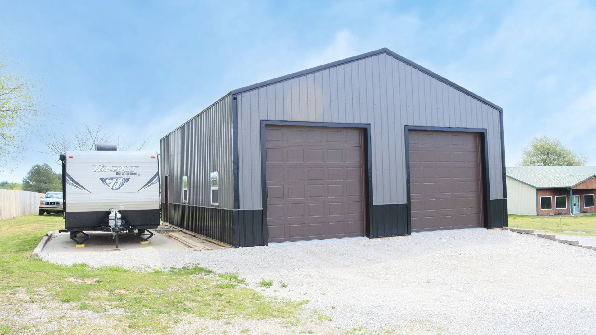 Gray metal warehouse with two brown roll-up doors, beside a white RV on a gravel lot under a blue sky