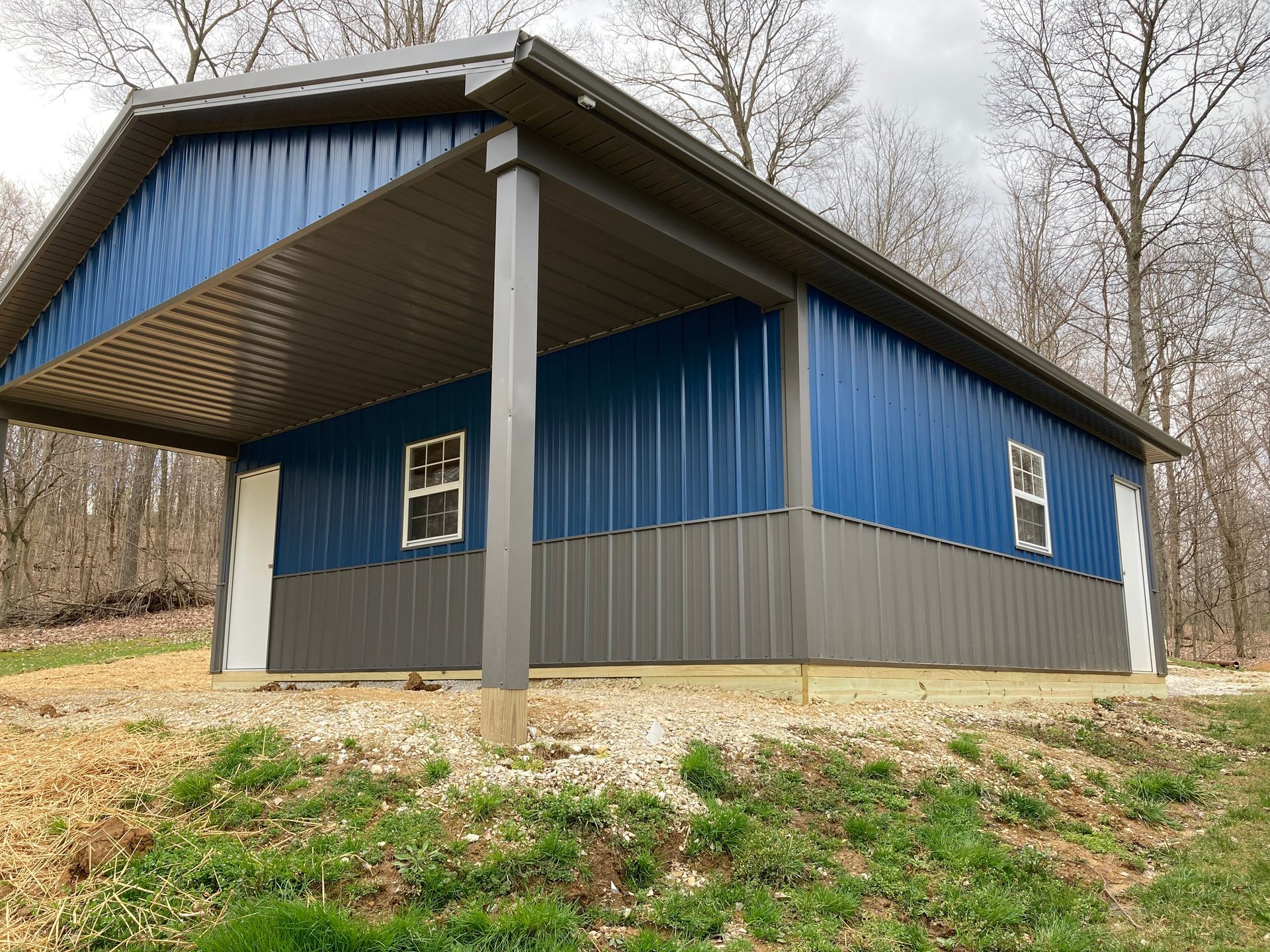 Blue metal barn-like building with a gray lower wall, seen on a wooded hillside.