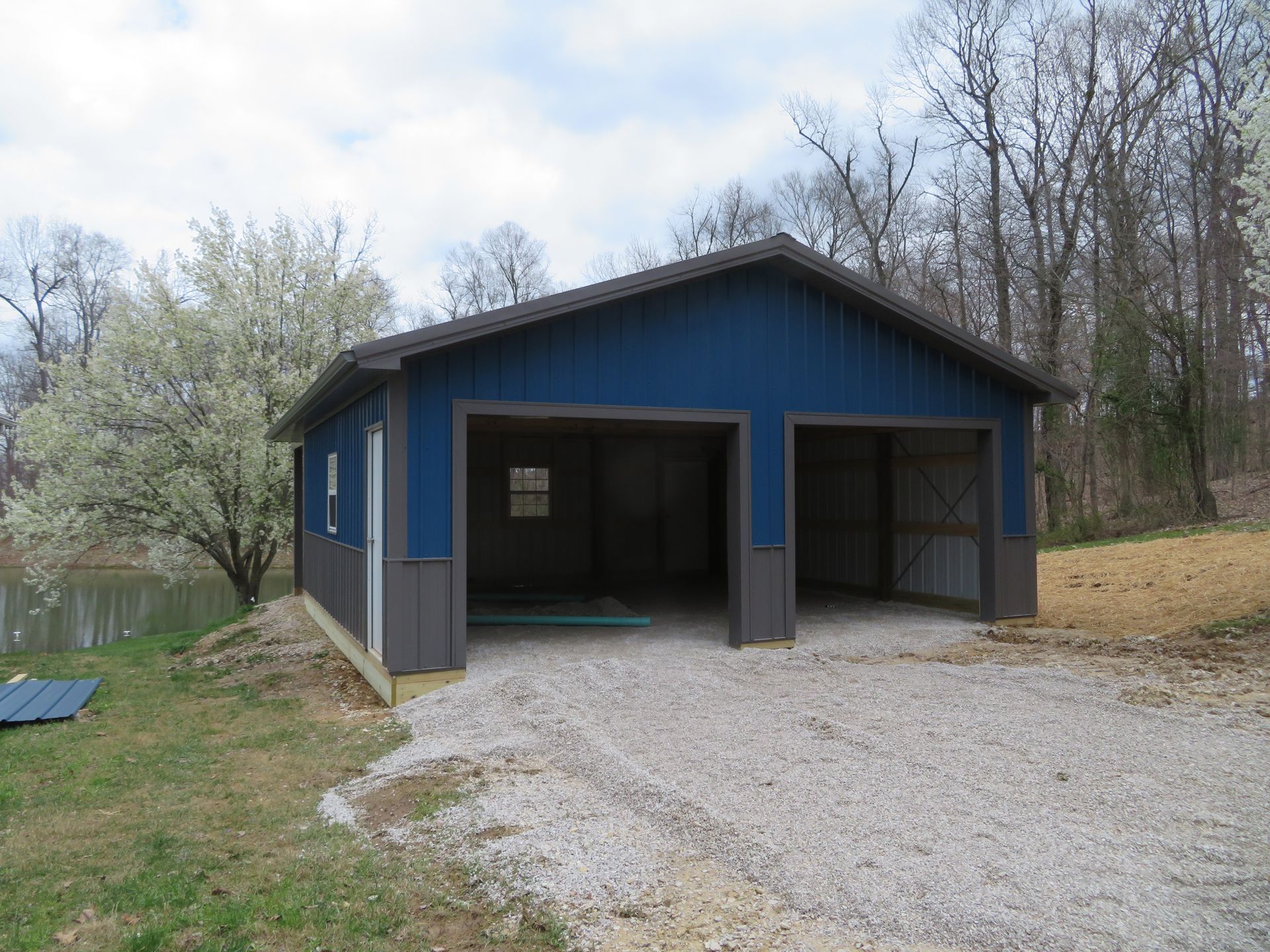 Blue detached garage with open bays and a gravel driveway in a wooded yard