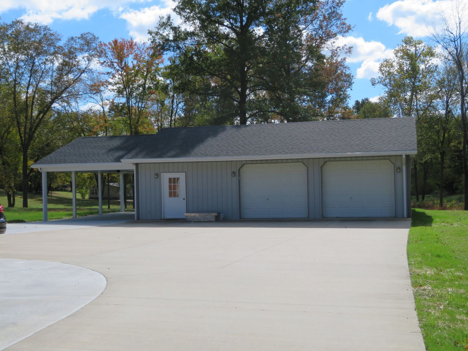 Gray garage with two doors and a carport beside a wide concrete driveway, surrounded by trees and grass.