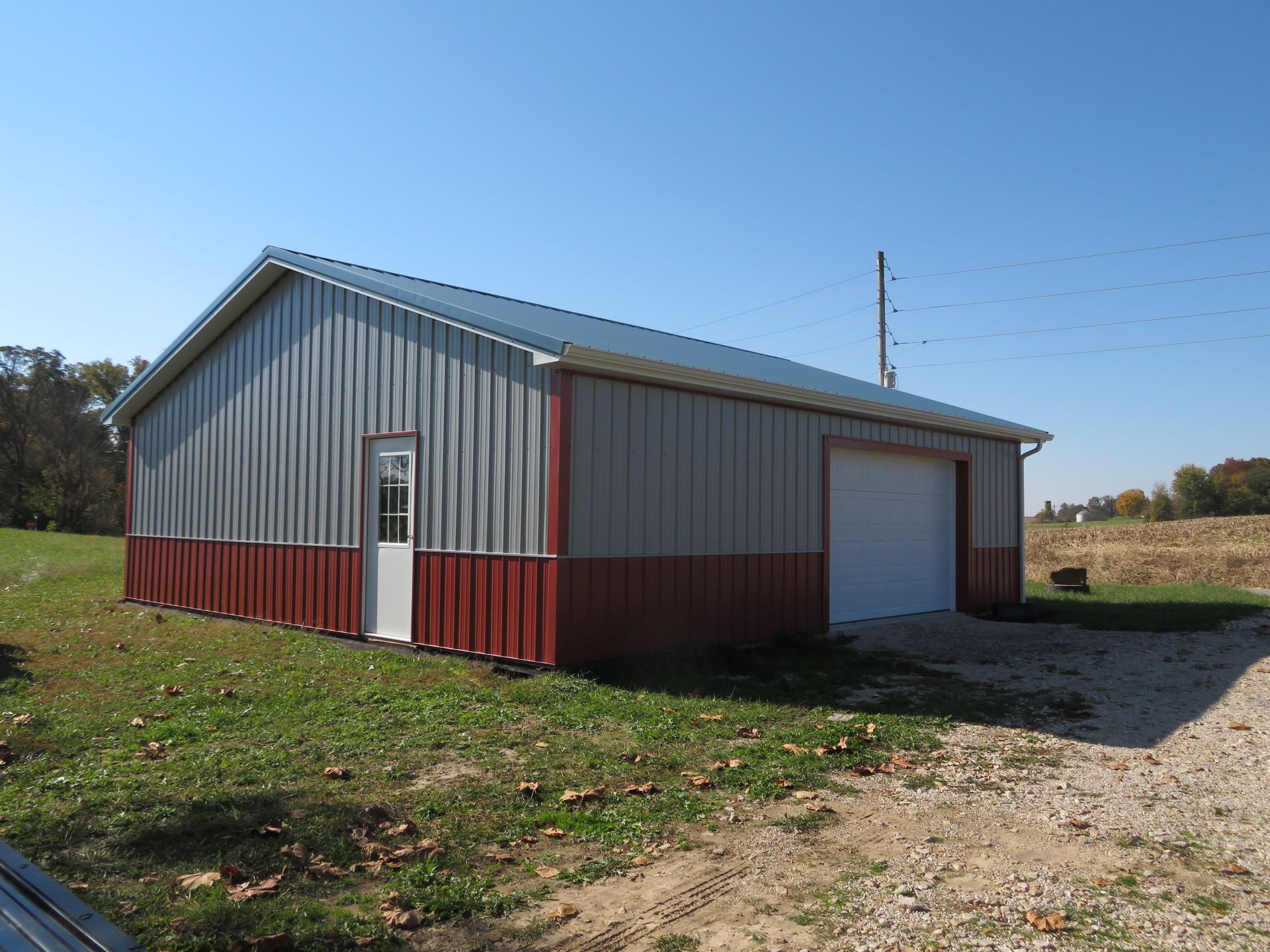 Red and gray metal barn-style building with a garage door beside a gravel drive on a sunny rural lot
