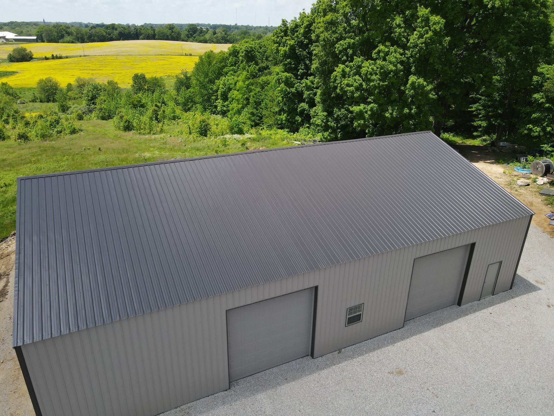 Gray metal storage building with three garage doors on a gravel lot, surrounded by green trees and fields.
