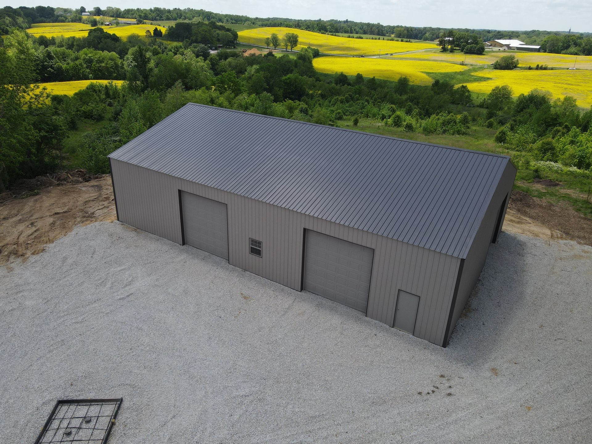 Gray metal storage building with a dark roof on a gravel lot, surrounded by green fields and trees