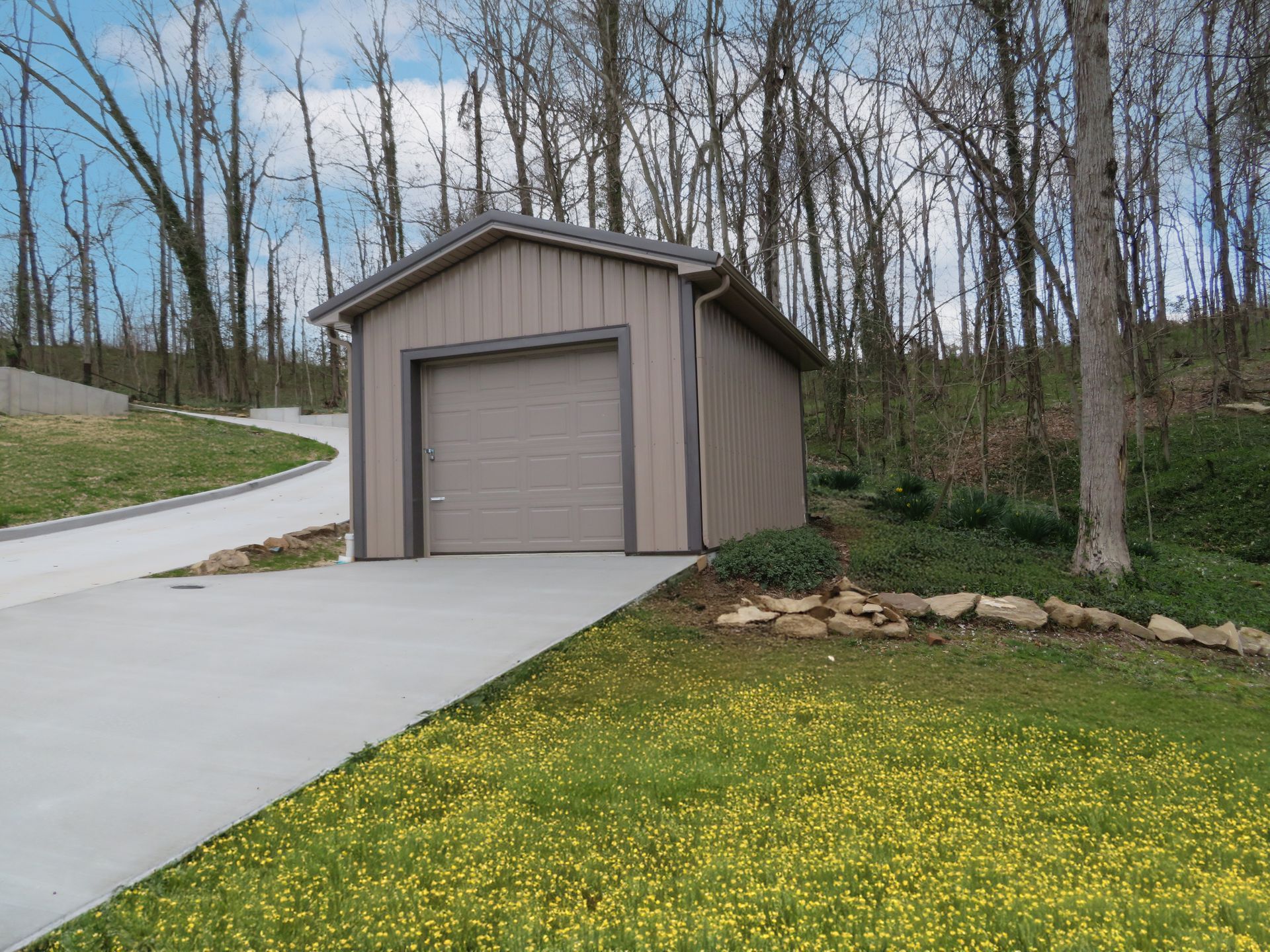 Small beige detached garage beside a concrete driveway, with trees and a grassy yard in the background.