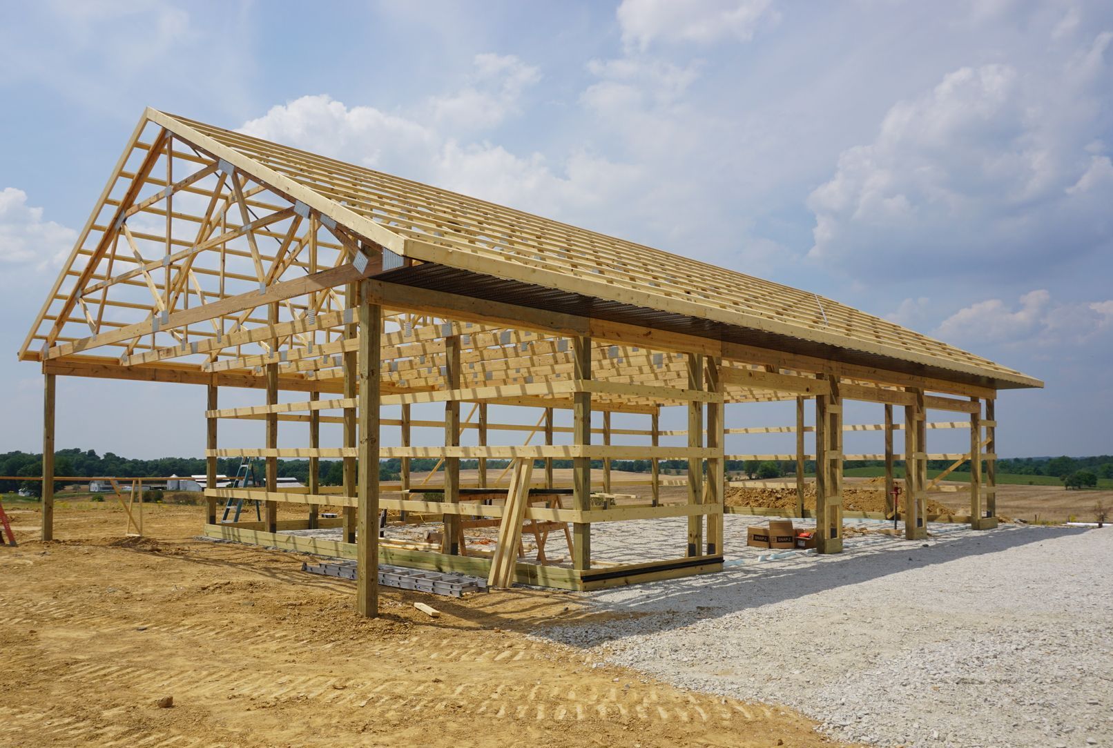 Wooden house frame under construction on a gravel lot under a partly cloudy sky