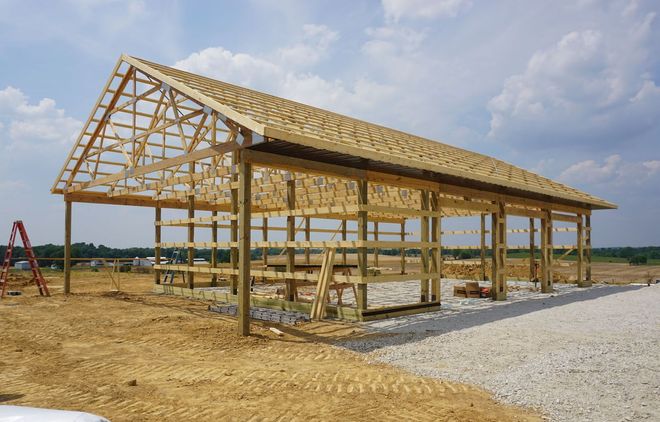 Partially framed wooden barn under construction on a dirt lot beside a gravel road