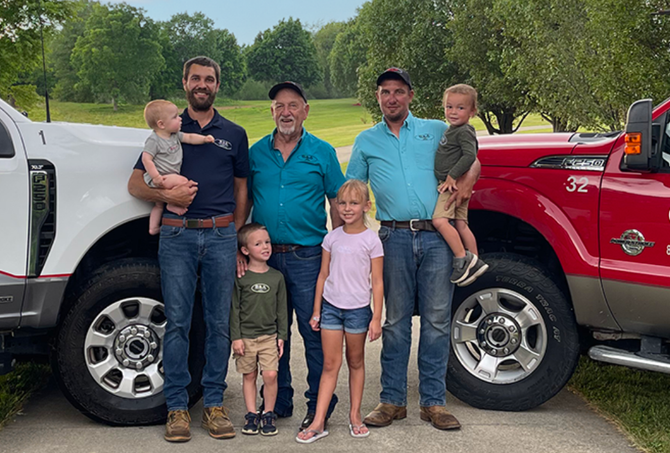 Family posing between white and red pickup trucks in a driveway, with two children and two small dogs
