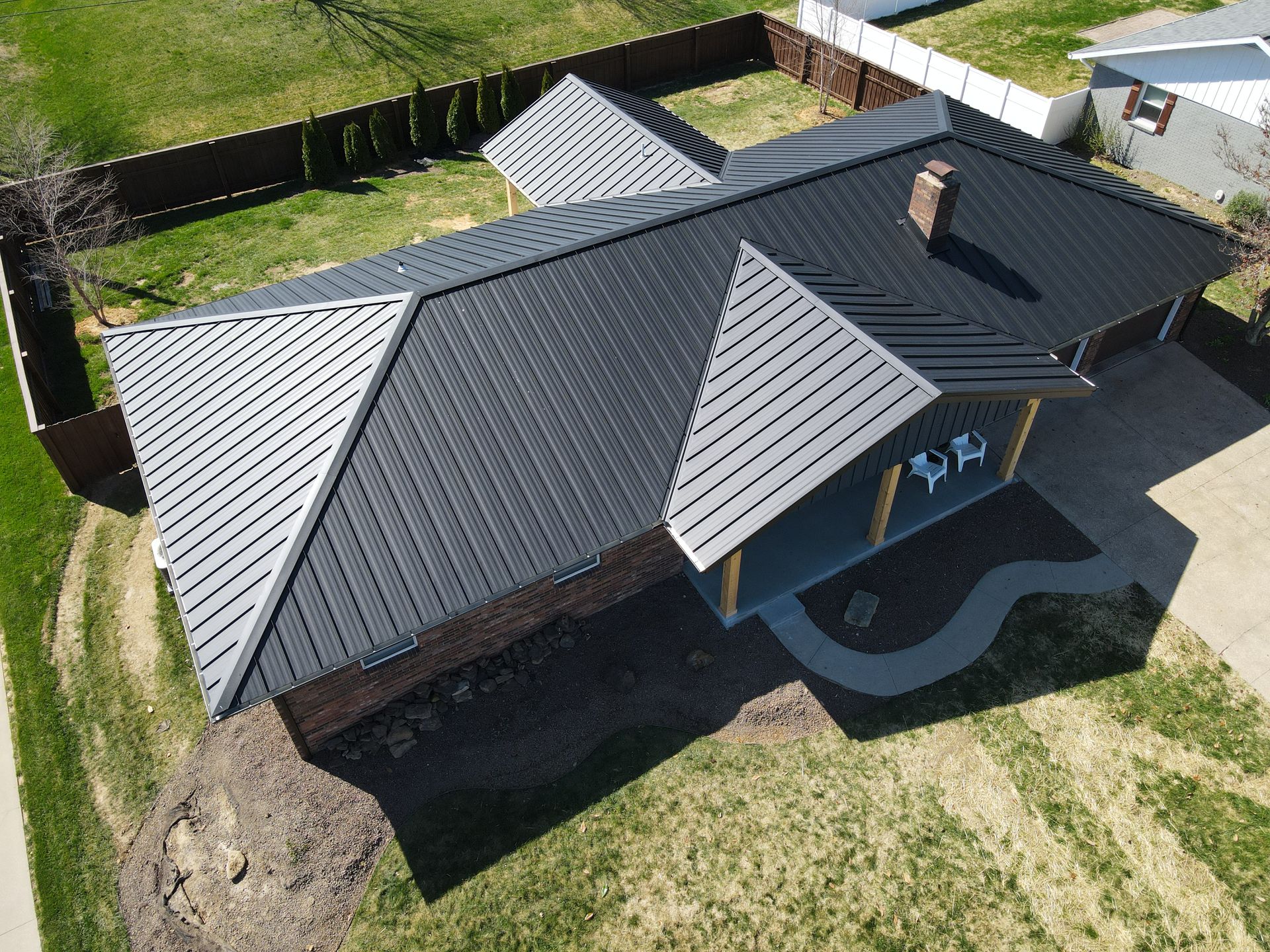 Aerial view of a house with a dark metal roof and small covered porch, surrounded by lawn and fencing