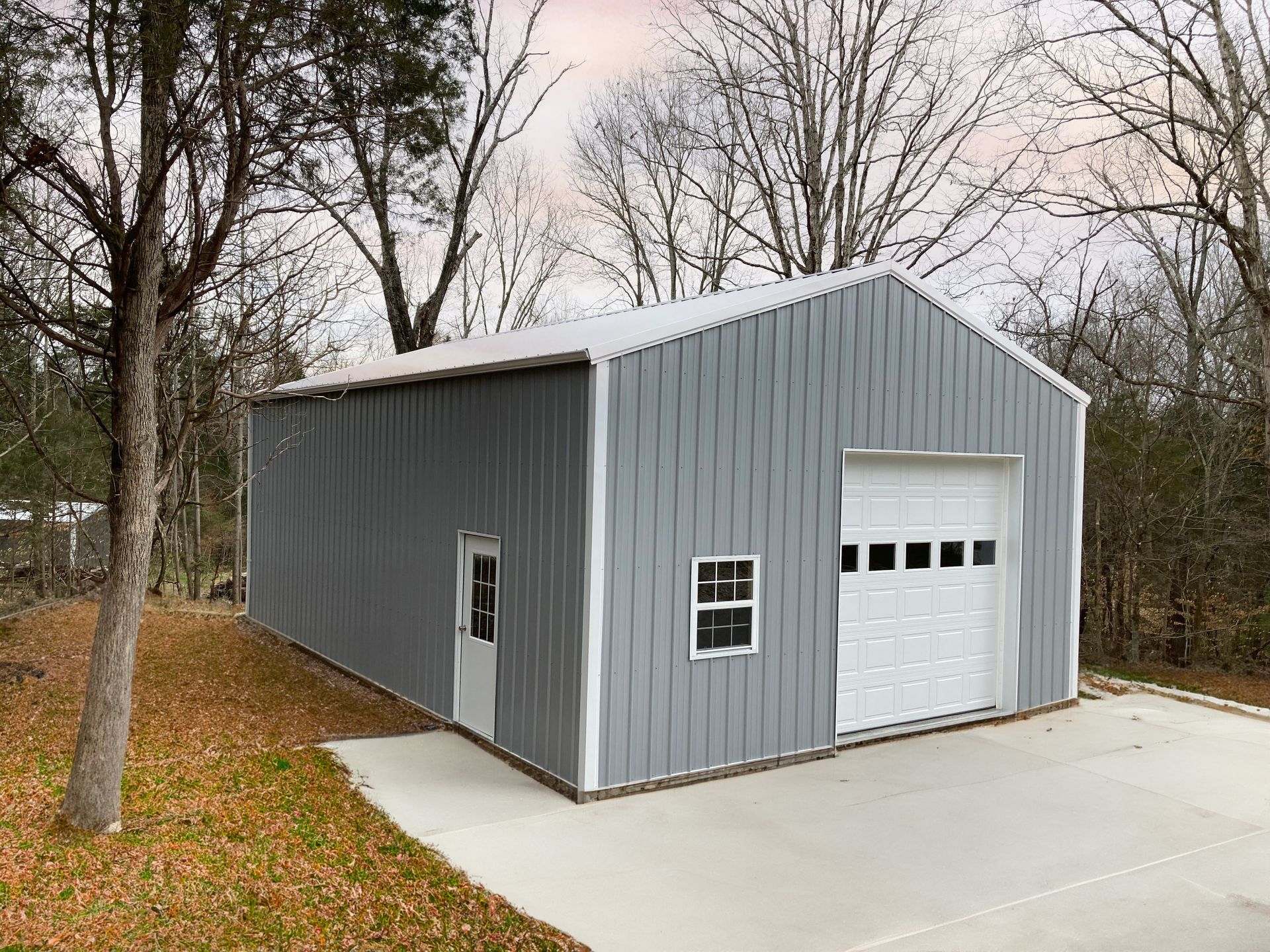Gray metal garage with a white roll-up door on a concrete pad, surrounded by leafless trees.