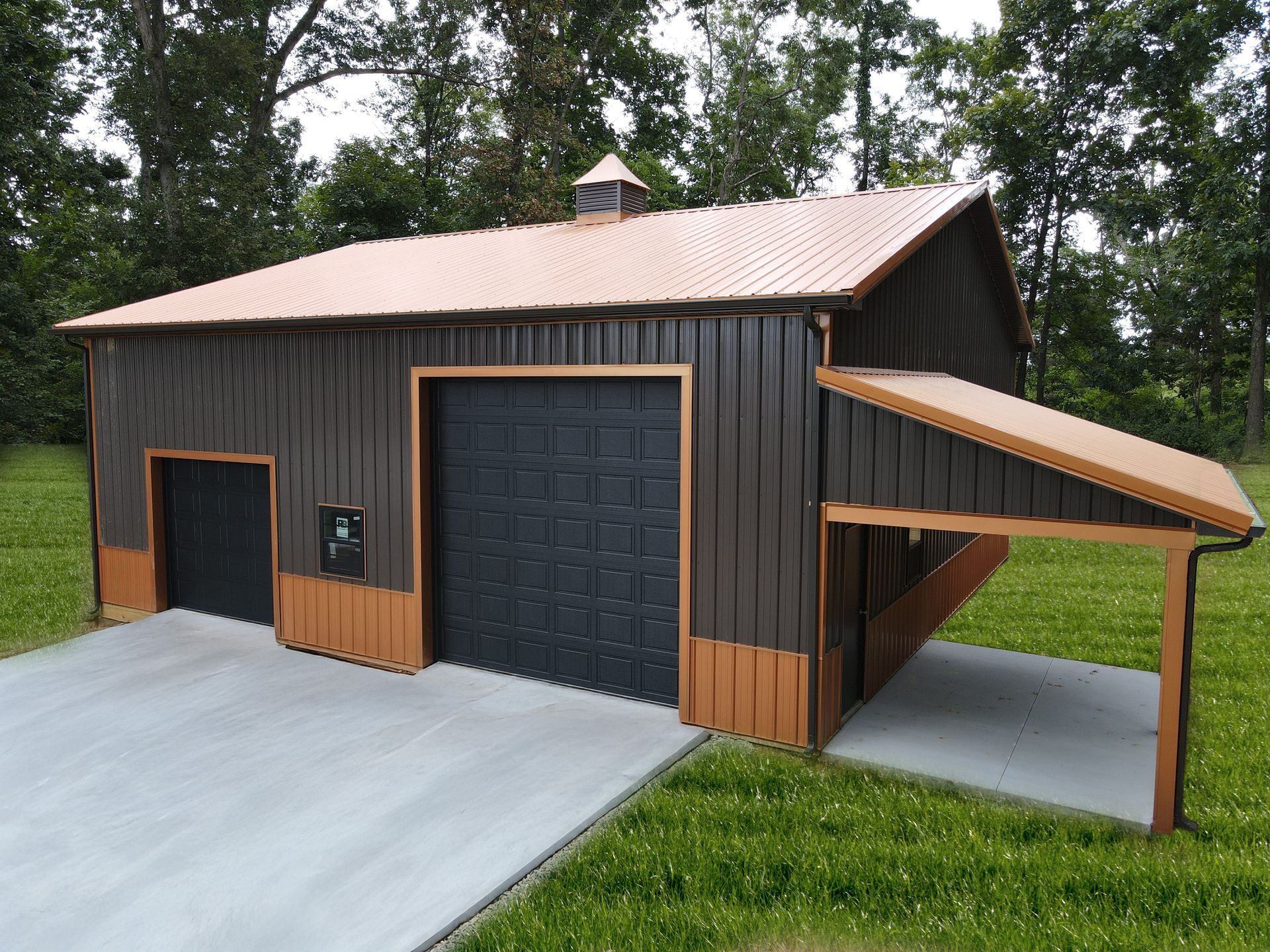 Dark brown barn-style garage with copper roof, large black doors, and a concrete driveway amid trees