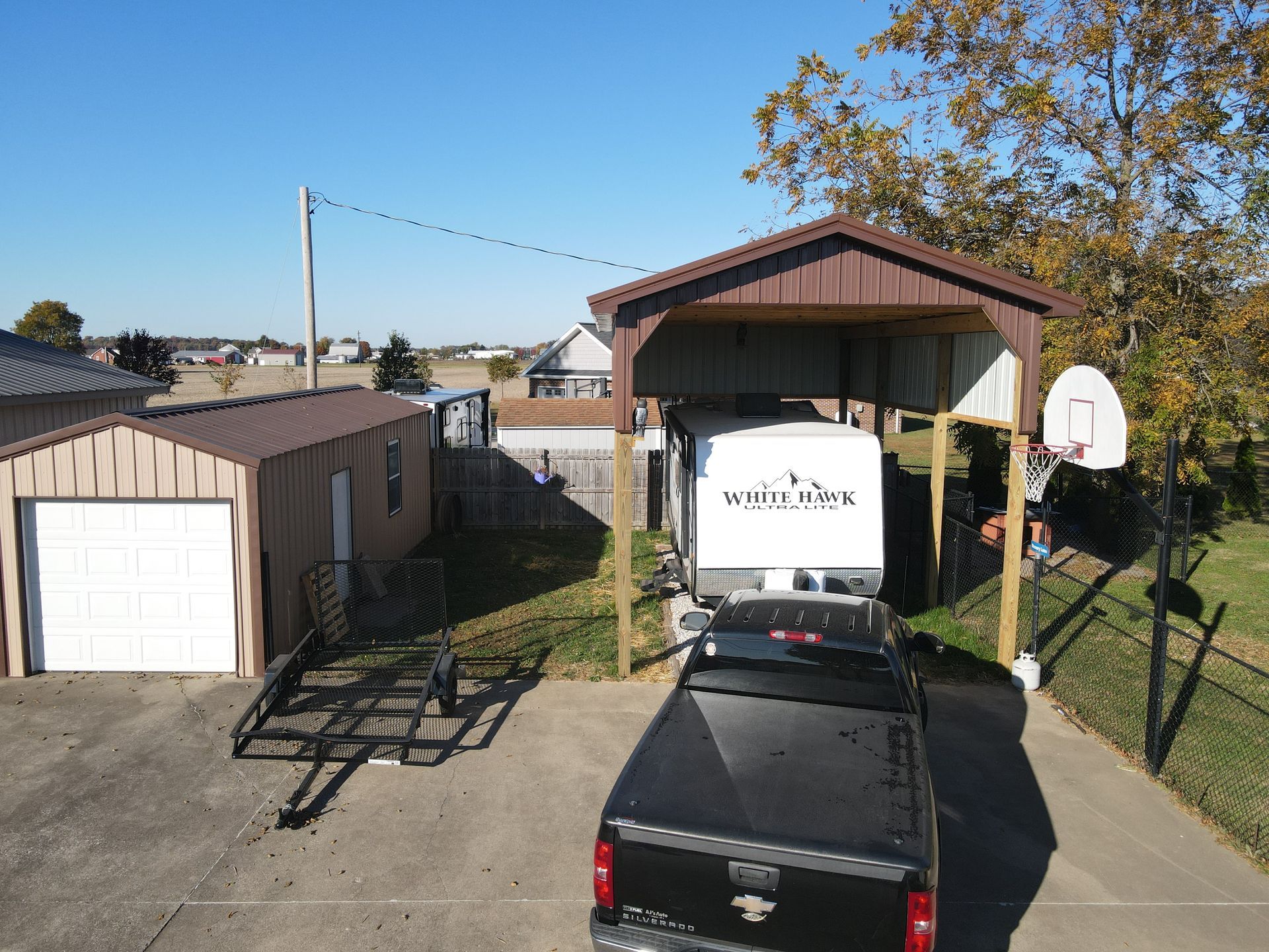 Driveway with black truck under a carport, white trailer, and detached garage on a sunny day