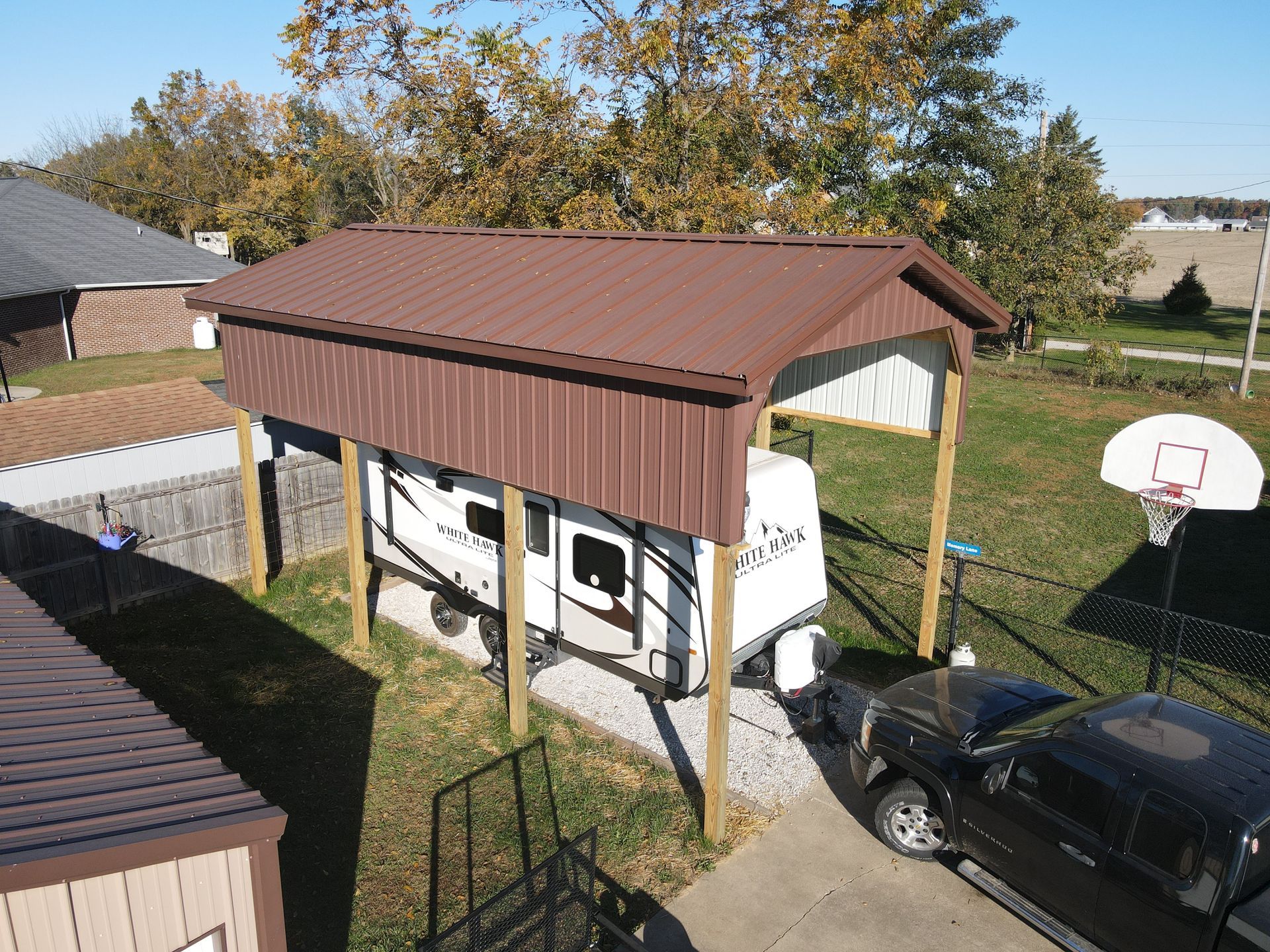 Carport with a white RV underneath, black car beside it, and a basketball hoop in a backyard.