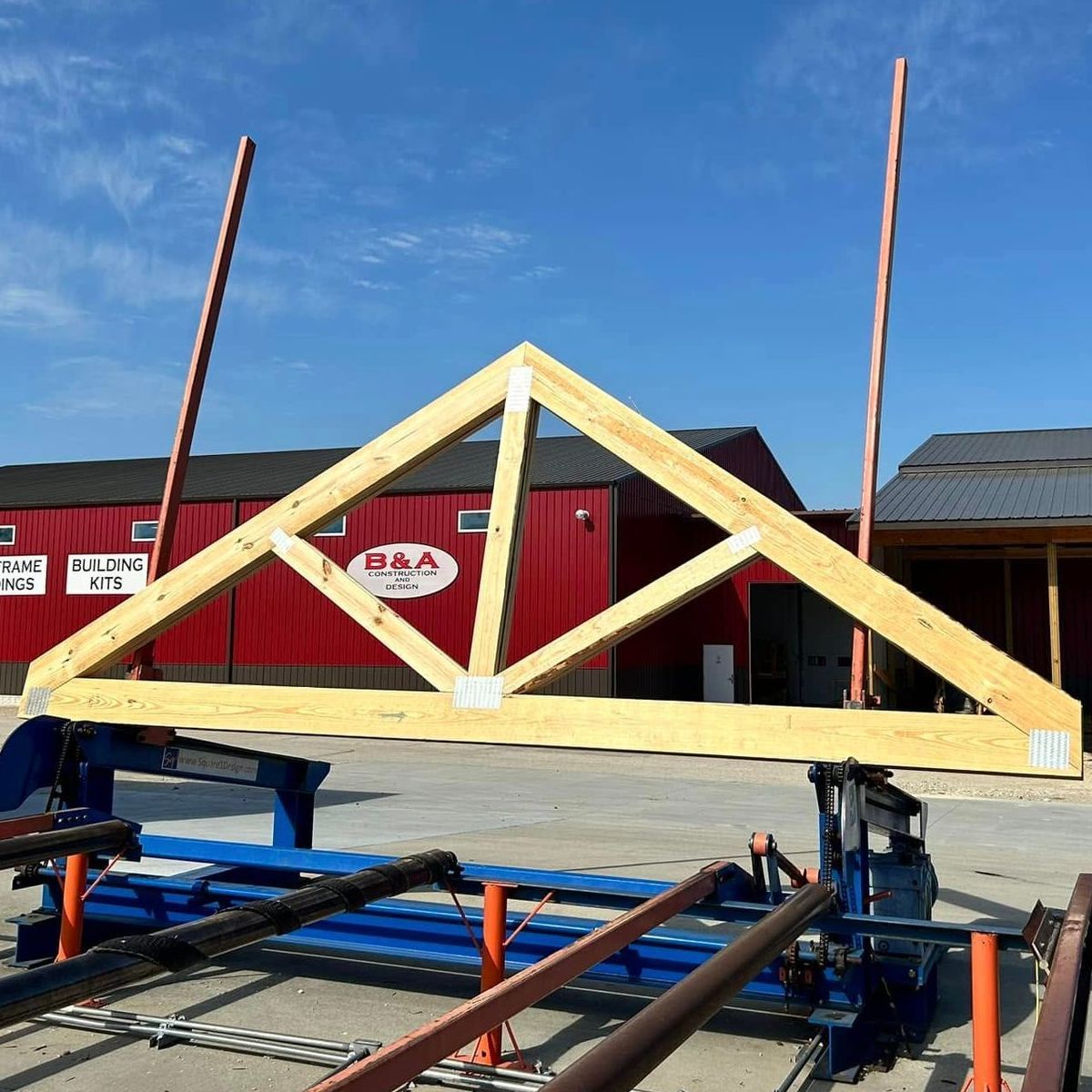 Wooden roof truss being built on a flatbed trailer outdoors under a blue sky.