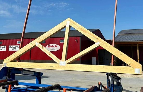 Wooden roof truss being lifted on a blue trailer, with a red building in the background.