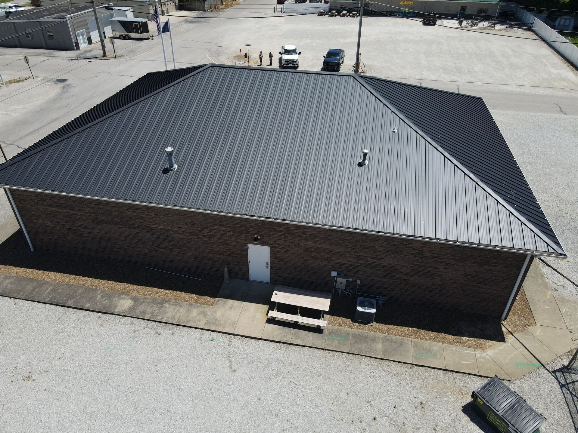 Aerial view of a small brick building with a dark metal roof and surrounding gravel lot.