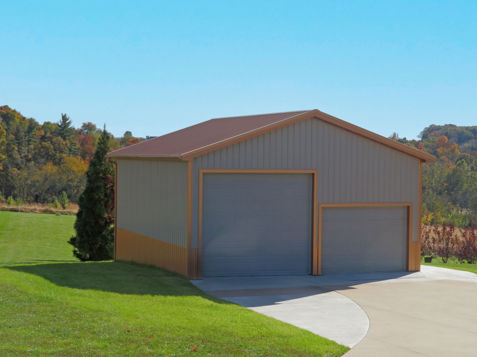 Metal garage building with tan siding and two doors beside a curved concrete driveway under a clear blue sky