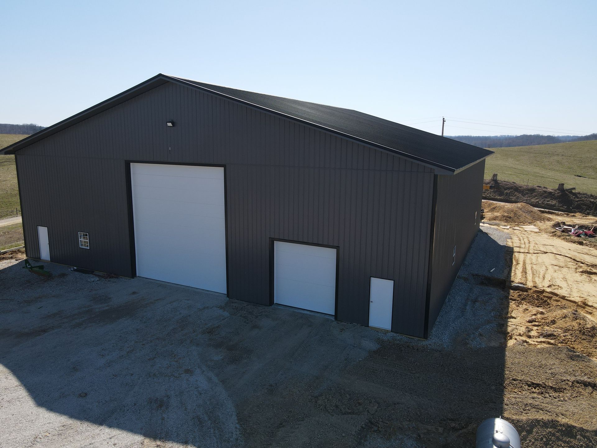 Dark gray metal barn with white doors on a gravel lot in a rural landscape