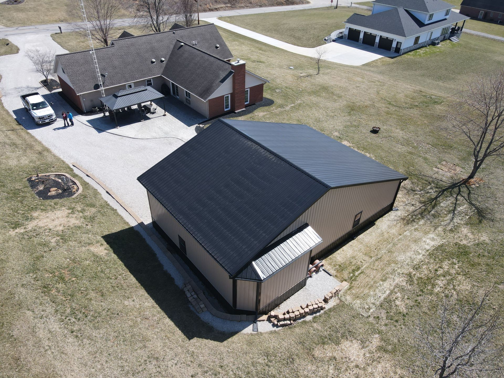 Aerial view of a house with a dark roof, driveway, and parked vehicles in a rural neighborhood