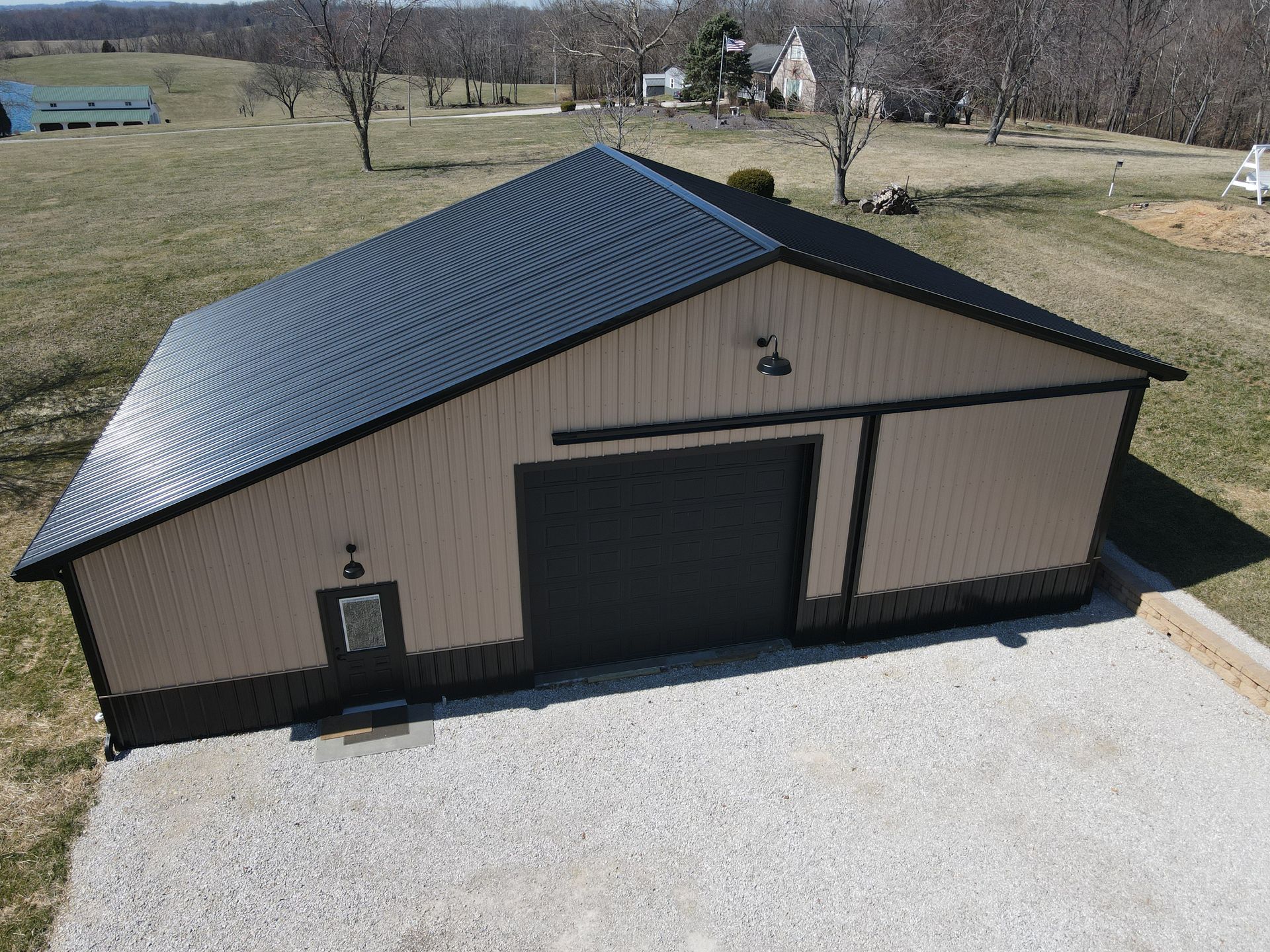 Tan metal building with black roof and large garage door on a gravel drive in a rural field