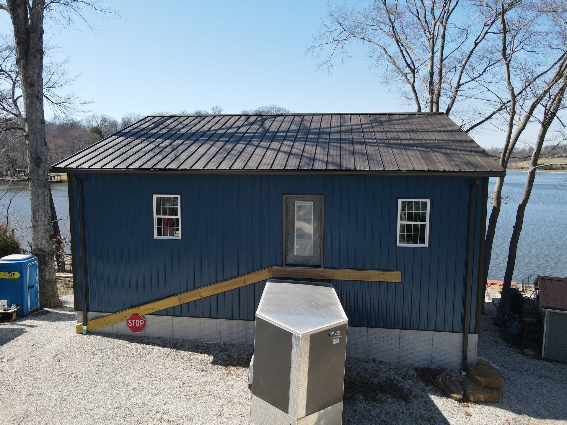 Blue lakeside cabin with a ramp and utility boxes in front, bare trees behind