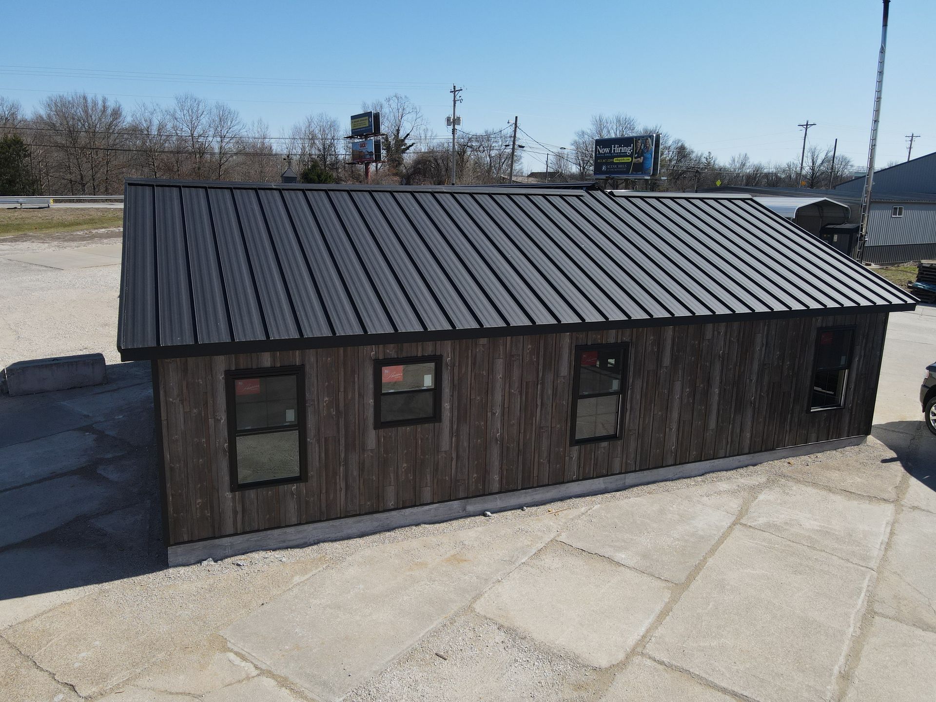 Small dark wooden building with a black metal roof on a paved lot under a clear sky.
