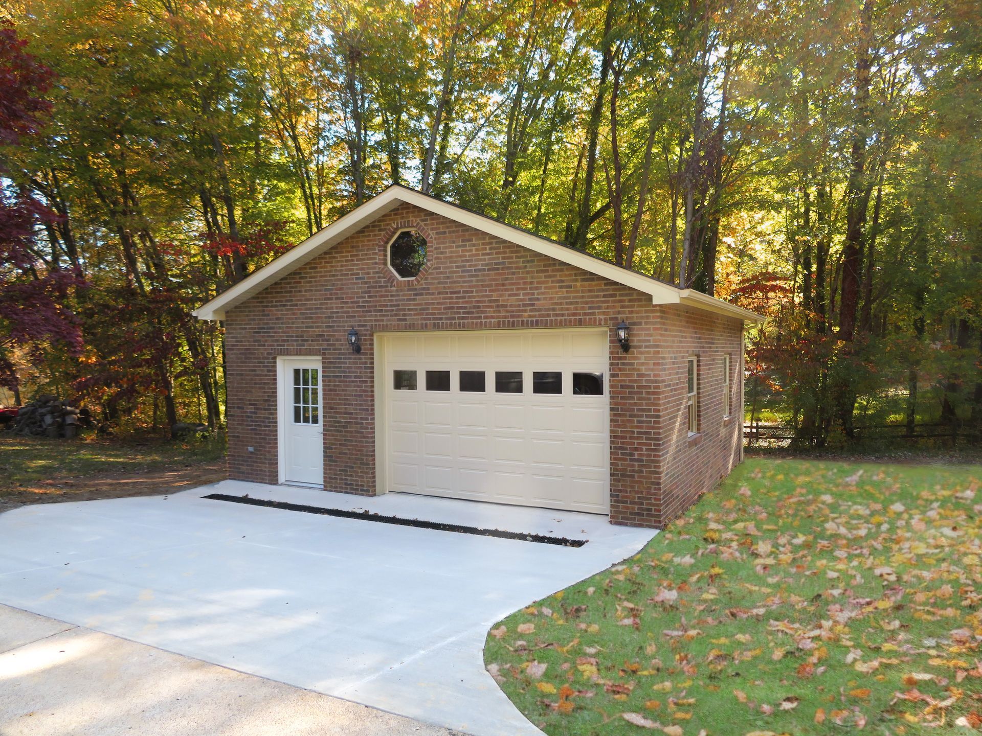 Detached brick garage with white door and concrete driveway beside a wooded yard in autumn