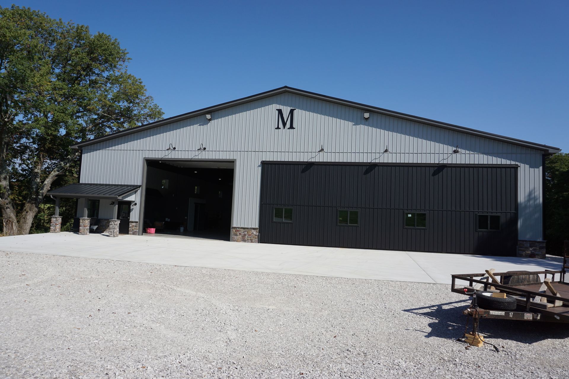 Large gray metal barn with open doors and a black side section on a gravel lot under a clear blue sky.