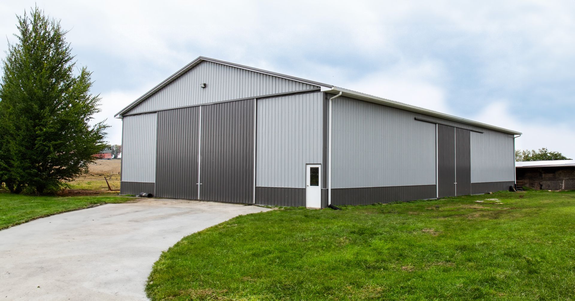 Gray metal barn on a grassy lot beside a curved concrete driveway under a cloudy sky