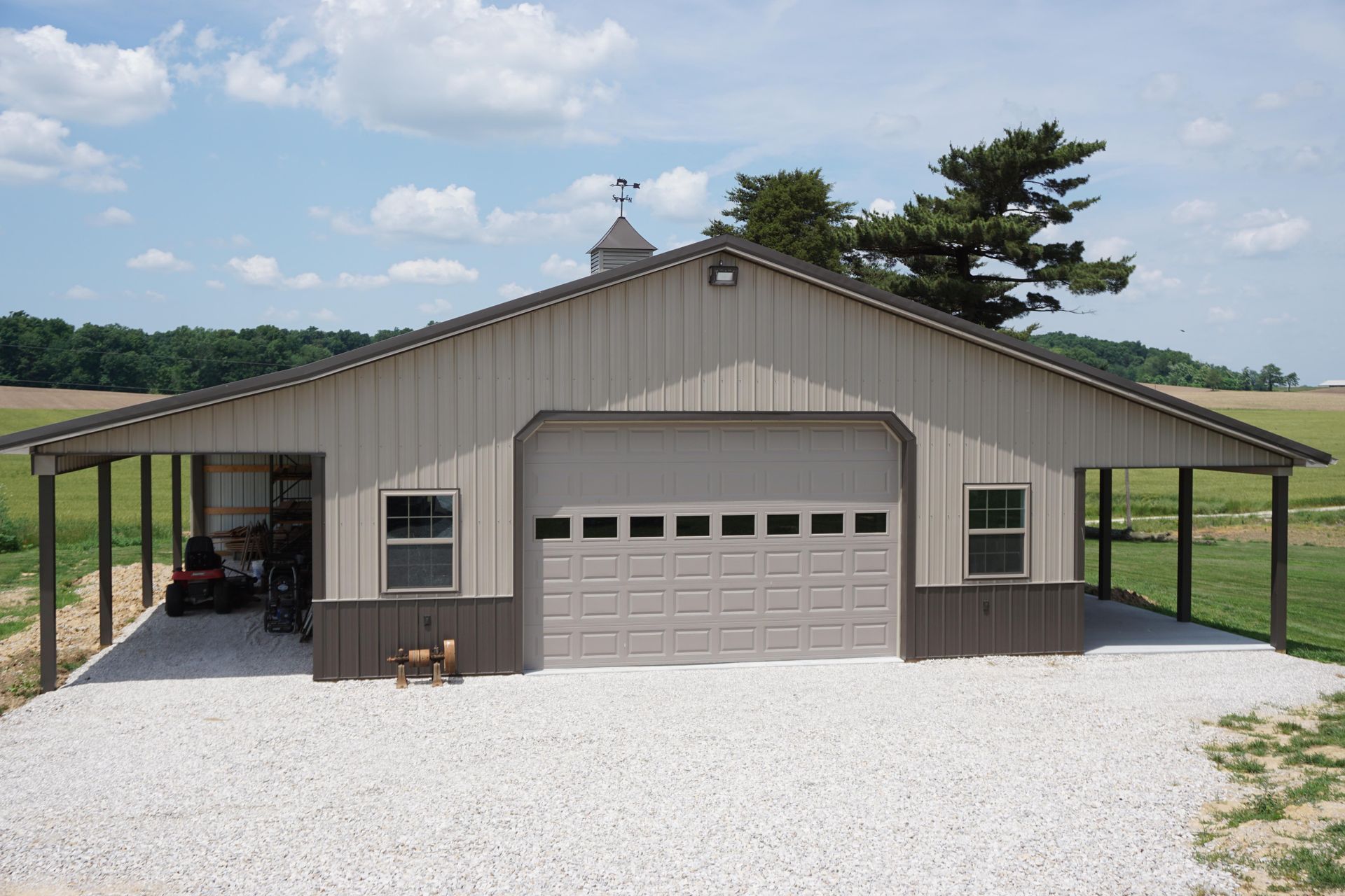 Large beige metal garage with white gravel driveway, side carport, and fields under a blue sky
