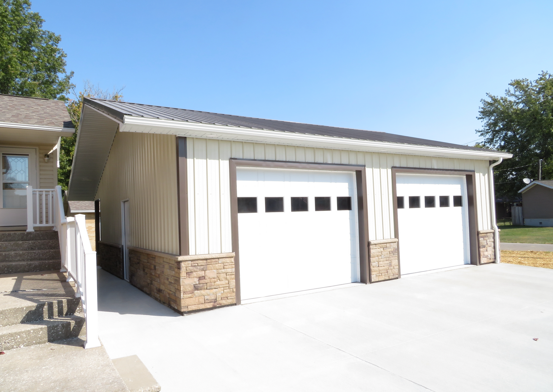 Detached two-car garage with beige siding, stone base, and white doors on a sunny driveway