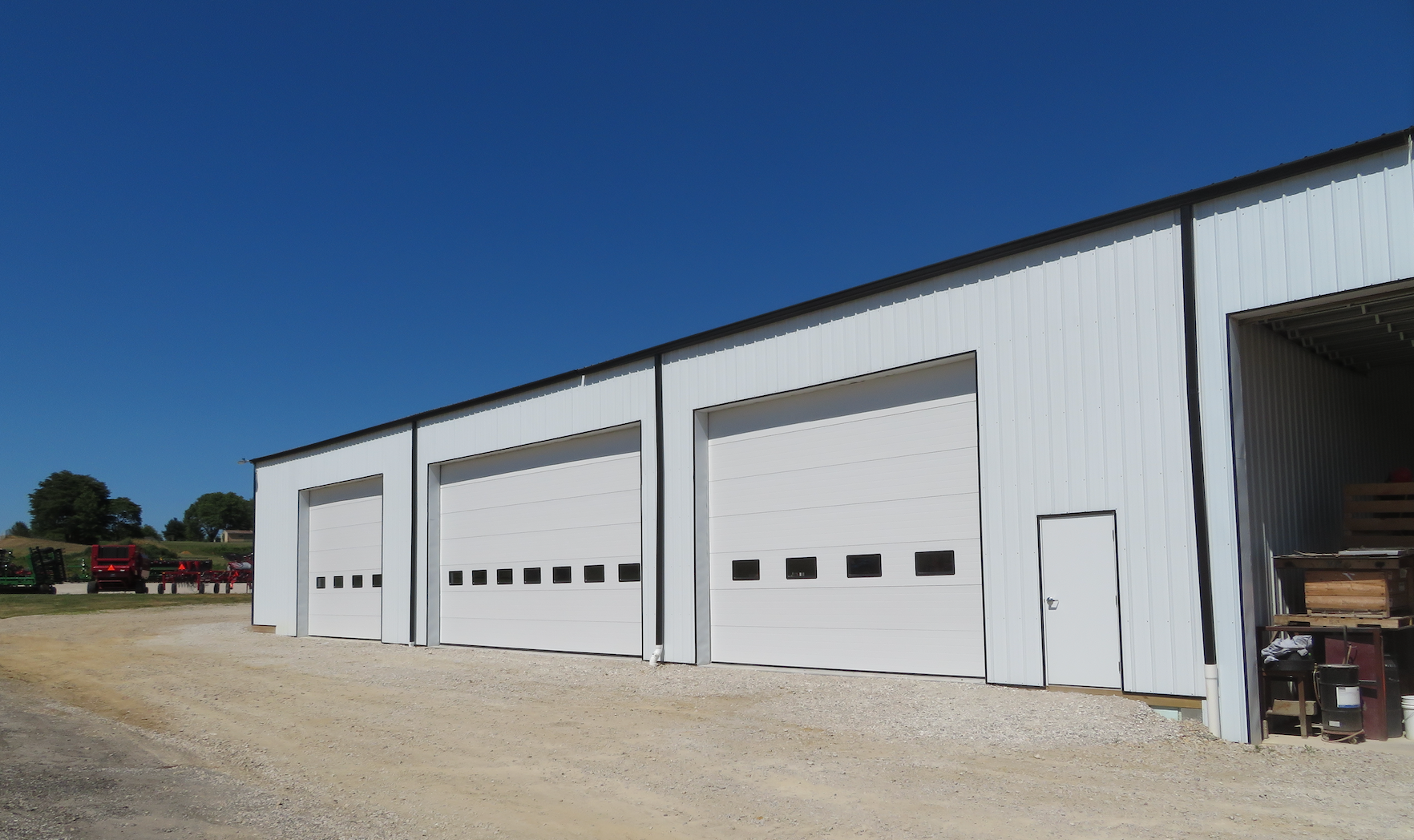 Large white industrial building with three garage bay doors on a gravel lot under a clear blue sky