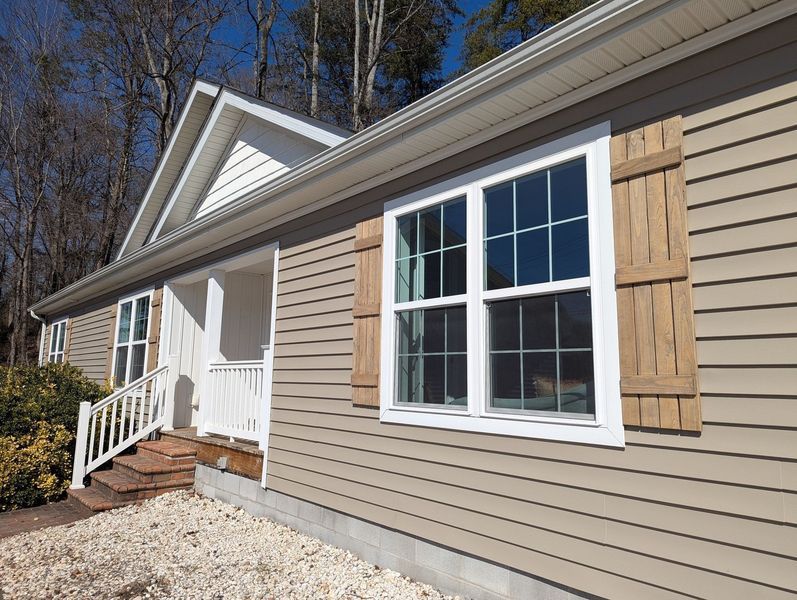 Tan house with light brown shutters, white window trim, and a porch, set against a backdrop of bare trees.