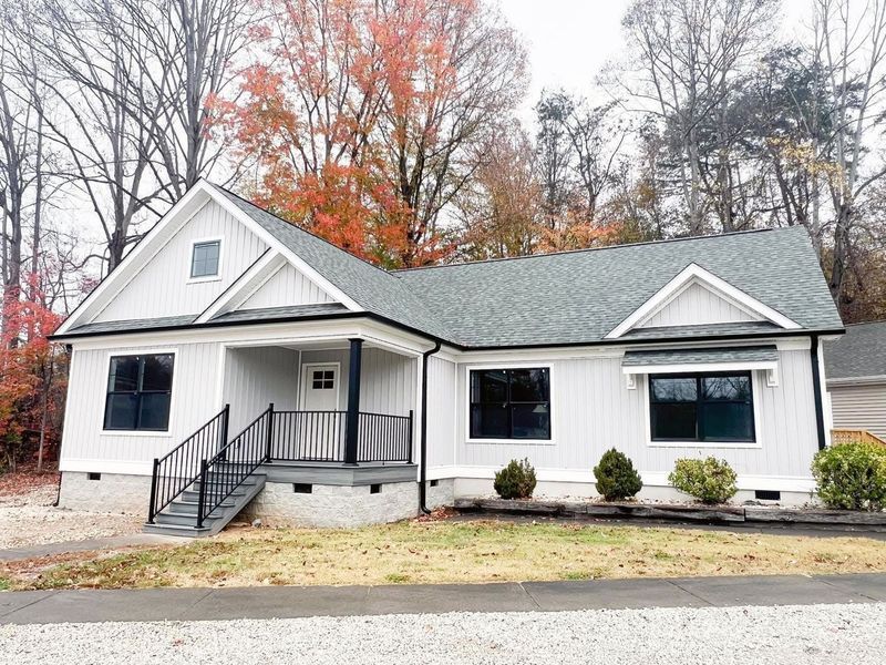 A one-story light gray farmhouse with a black metal roof, dark trim, and a small front porch, surrounded by autumn trees.
