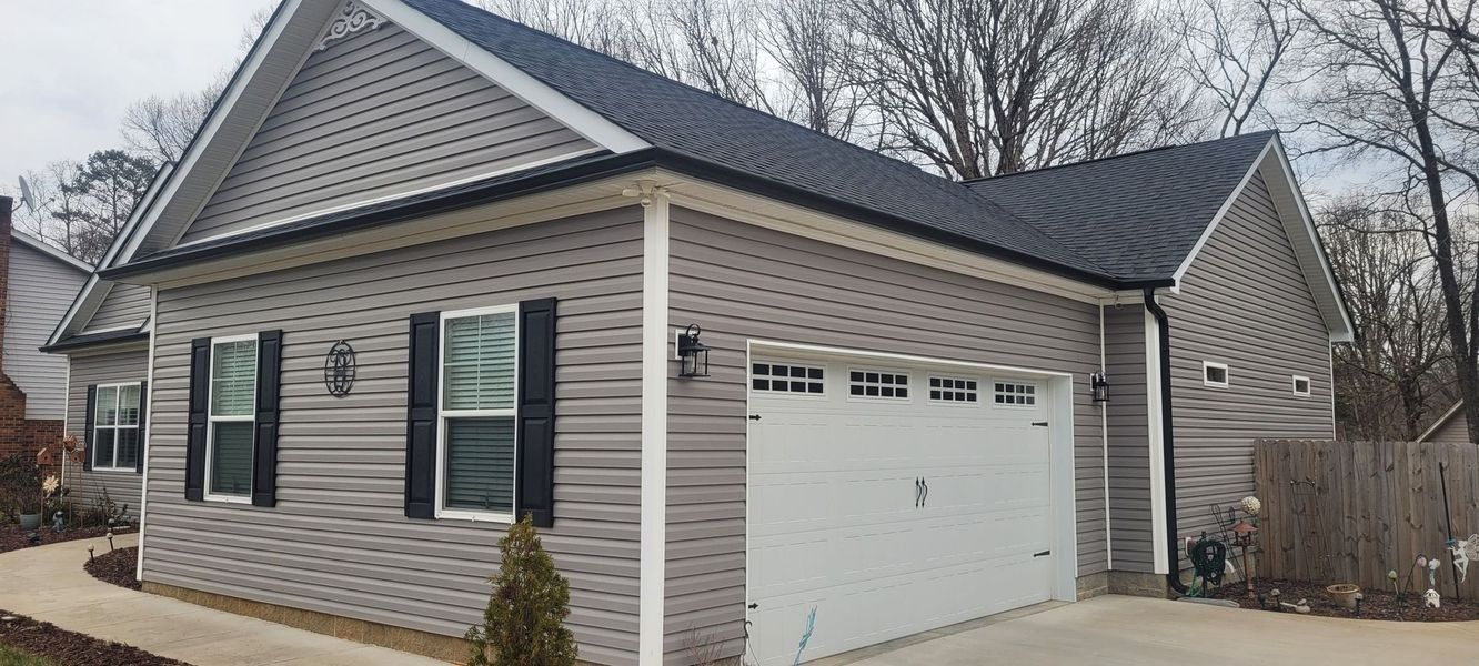 A gray vinyl-sided house with a black metal roof, white garage door, and a concrete walkway on a cloudy day.