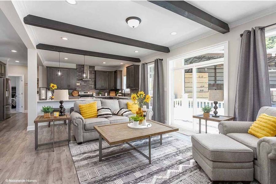 A living room with a beige sofa, gray armchair, patterned rug, wooden coffee table, and dark ceiling beams, near a kitchen.